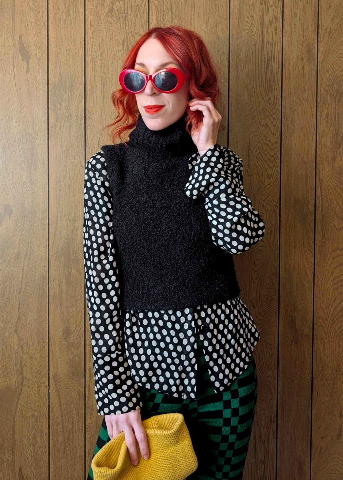 Woman wearing a black sleeveless turtleneck knit vest and  polka dot top underneath with a wooden wall background