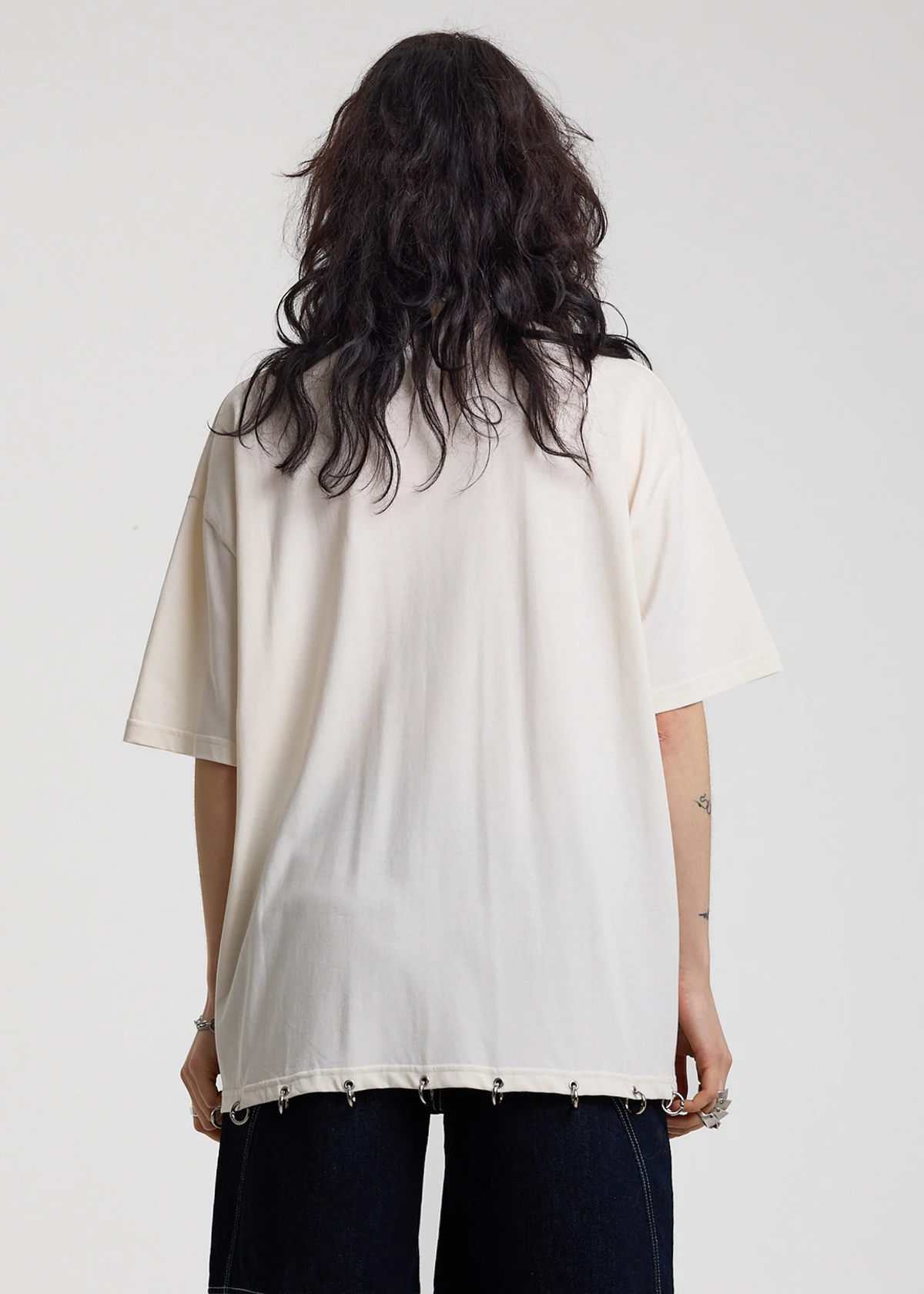 Back view of a person wearing an ivory t-shirt with silver O-ring details along the hem