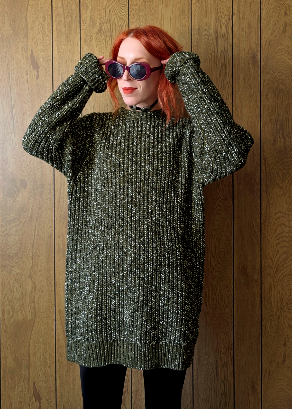 Person wearing an olive green chunky tunic sweater and sunglasses against a wooden background