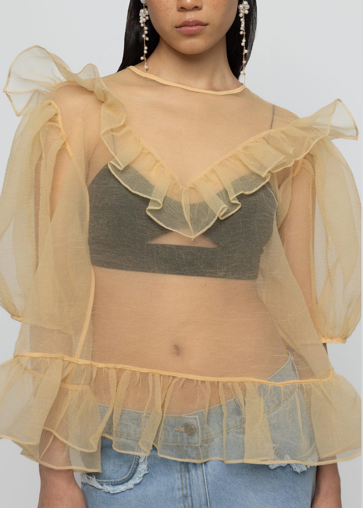 Woman wearing a sheer yellow organza balloon sleeve top over a dark bralette with denim jeans on a white background