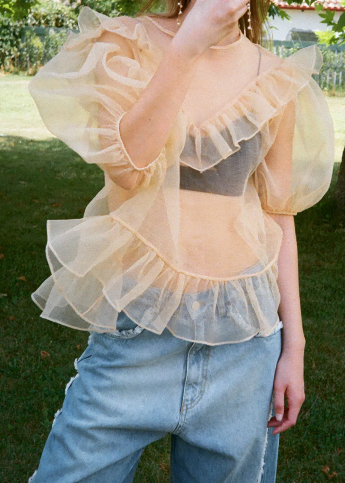 Person wearing a sheer ruffled top over a black bralette and blue jeans outdoors.