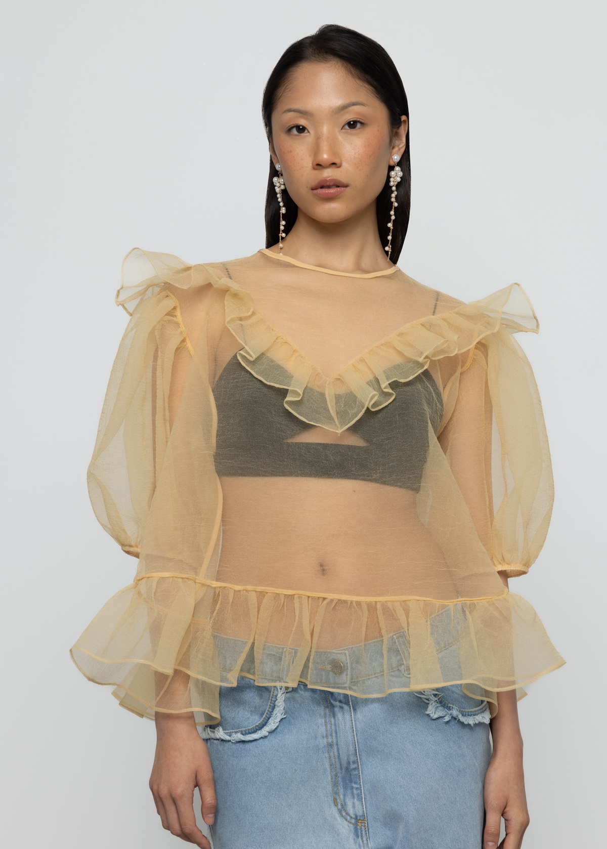 Woman wearing a sheer yellow organza balloon sleeve top over a dark bralette with denim jeans on a white background