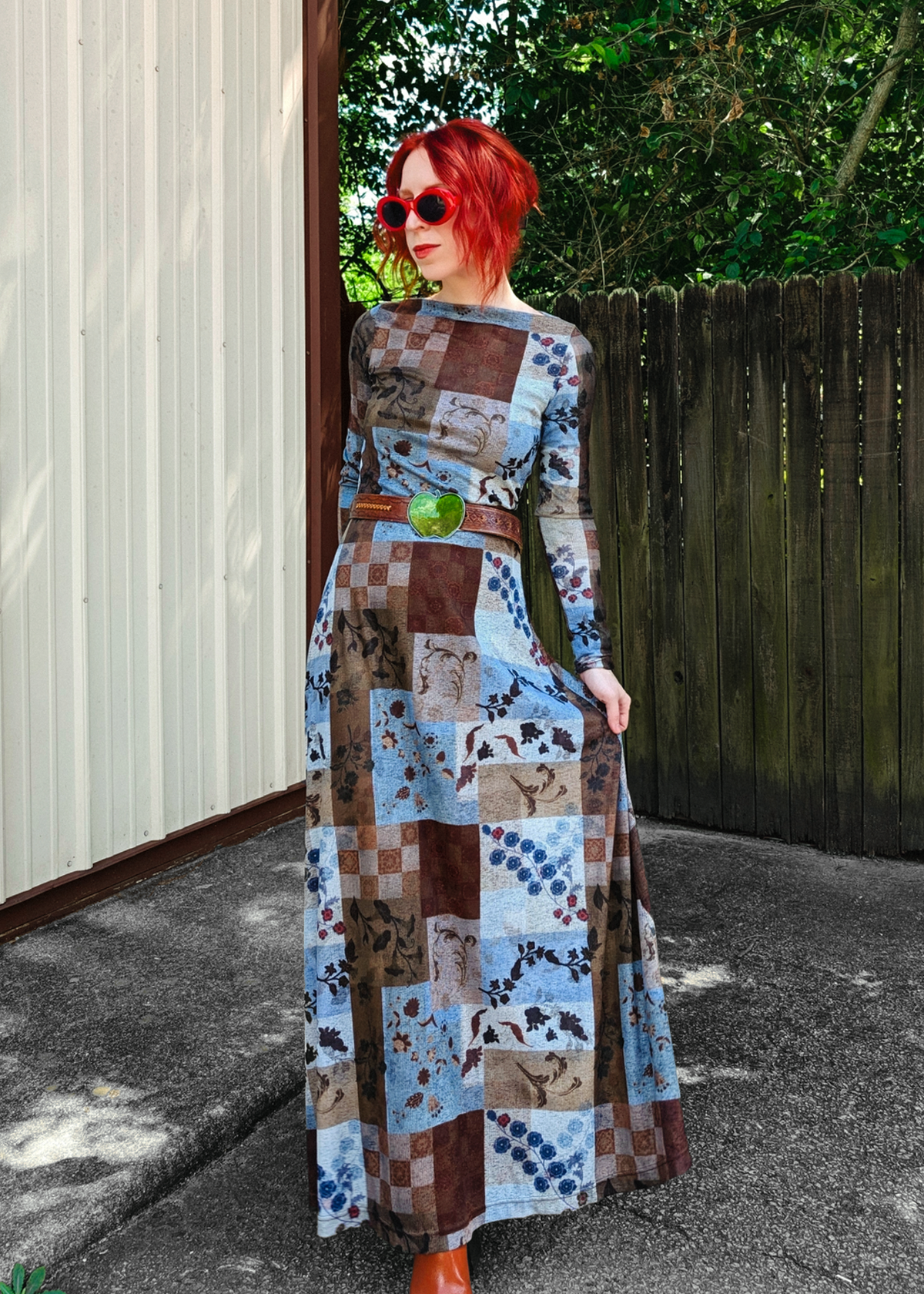 A woman with bright red hair and red sunglasses stands outdoors wearing a long-sleeve patchwork print maxi dress with blue, brown, and beige floral and checkered squares, paired with a brown belt and orange shoes.