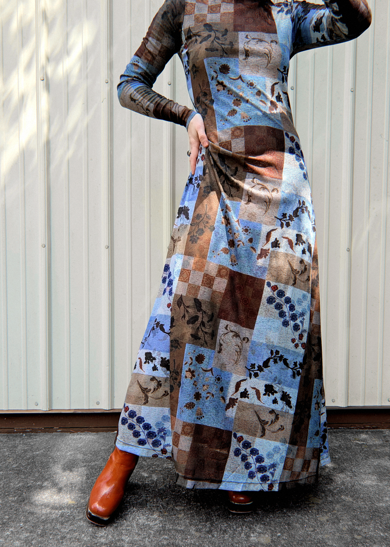 A woman wearing a long-sleeve patchwork print maxi dress with blue, brown, and beige tones, paired with orange leather ankle boots, standing in front of a white corrugated metal wall.