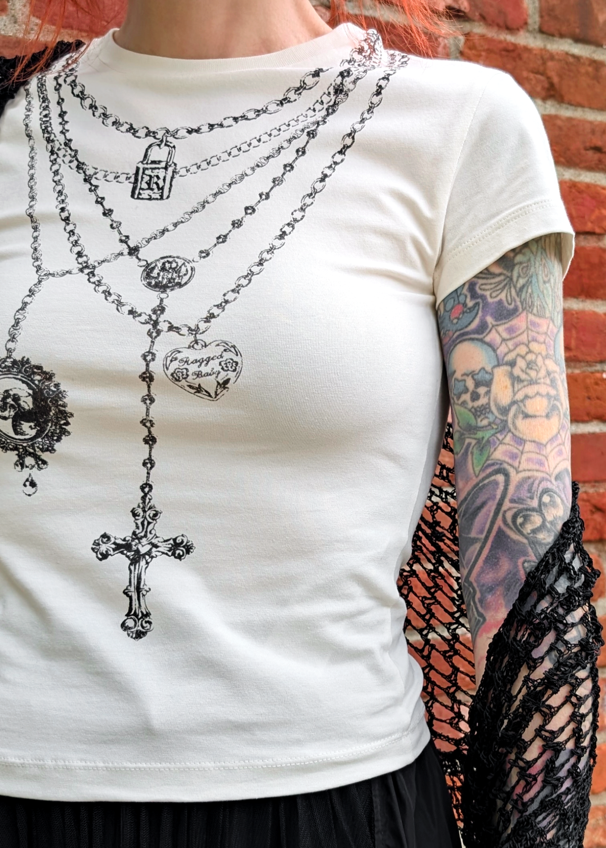 Person wearing an ivory short sleeve  top with graphic design of layered necklaces, styled with a crochet shawl and black skirt