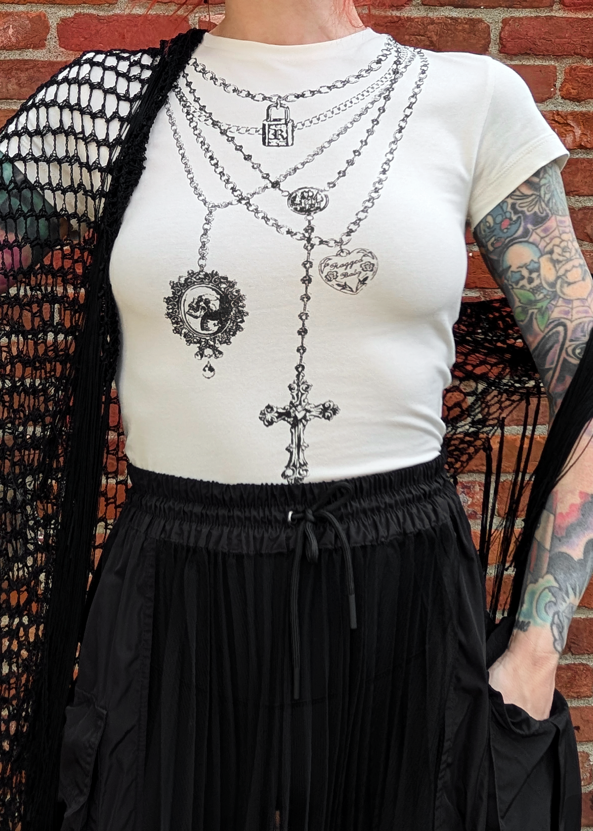 Person wearing an ivory short sleeve top with black graphic design of layered necklaces, styled with a black skirt and crochet shawl, standing against a brick wall.