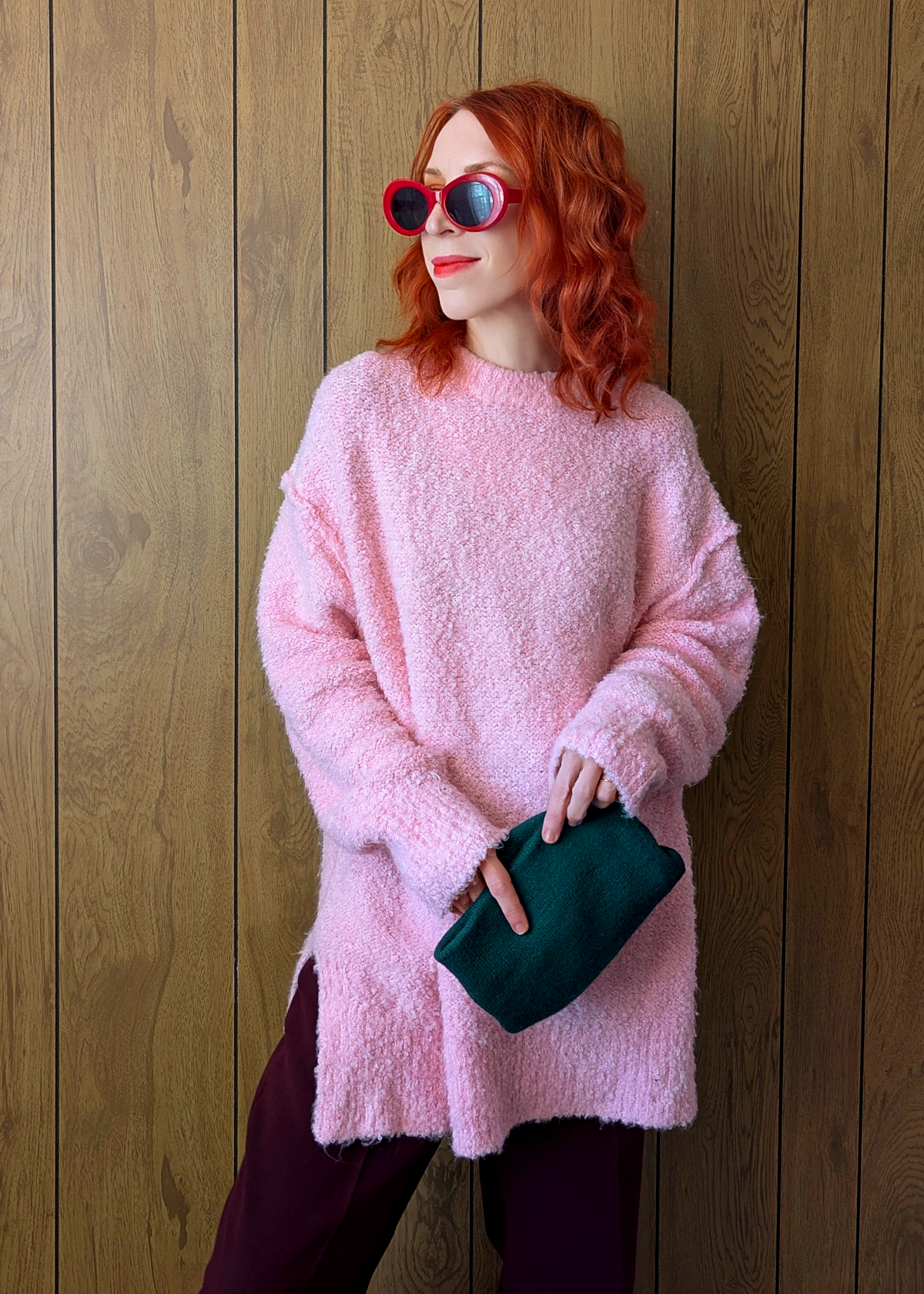 Woman wearing a pink sweater and sunglasses, holding a green clutch against a wooden wall.