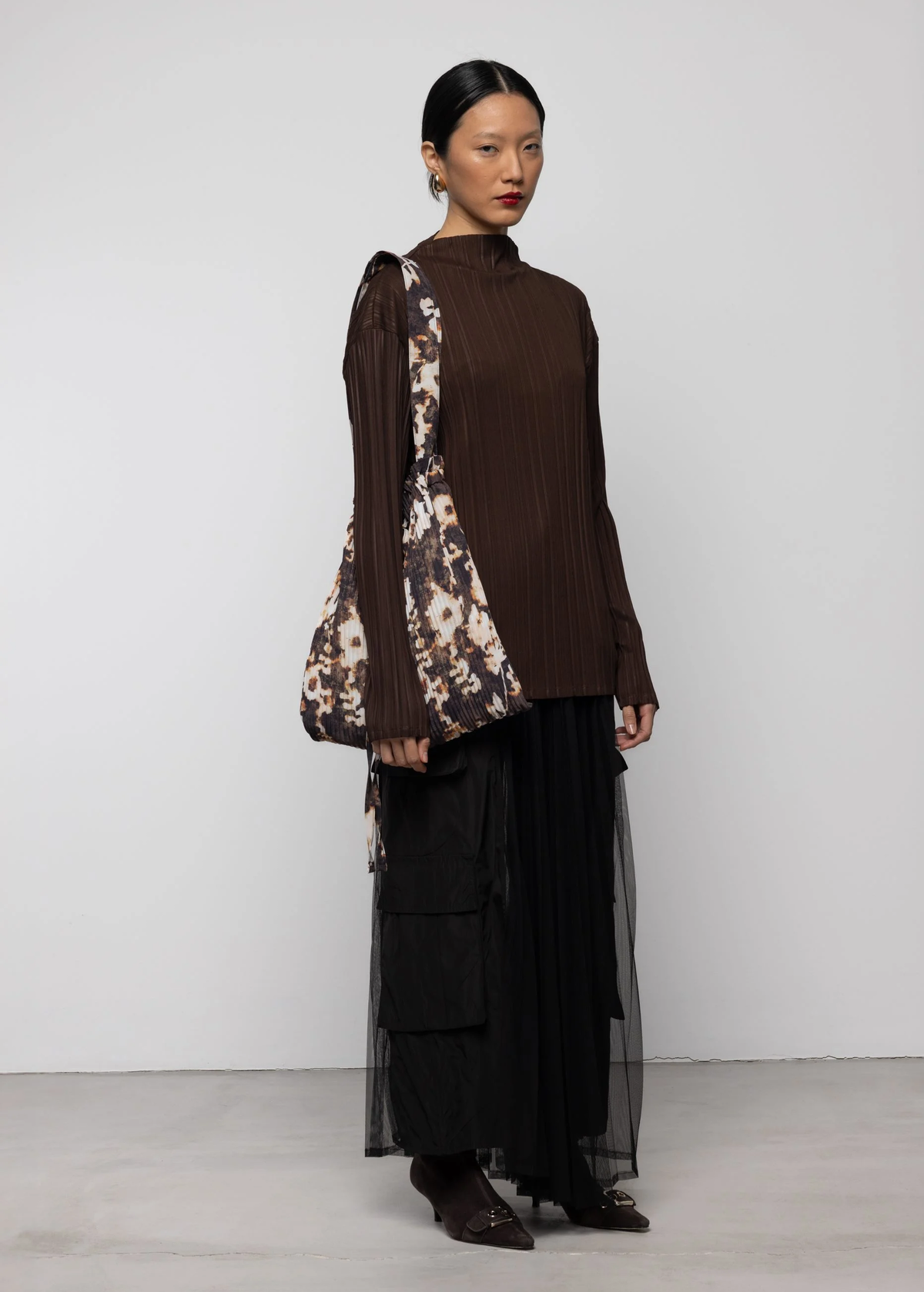 Woman wearing a brown top and black sheer mesh cargo skirt with a floral patterned bag against a white background