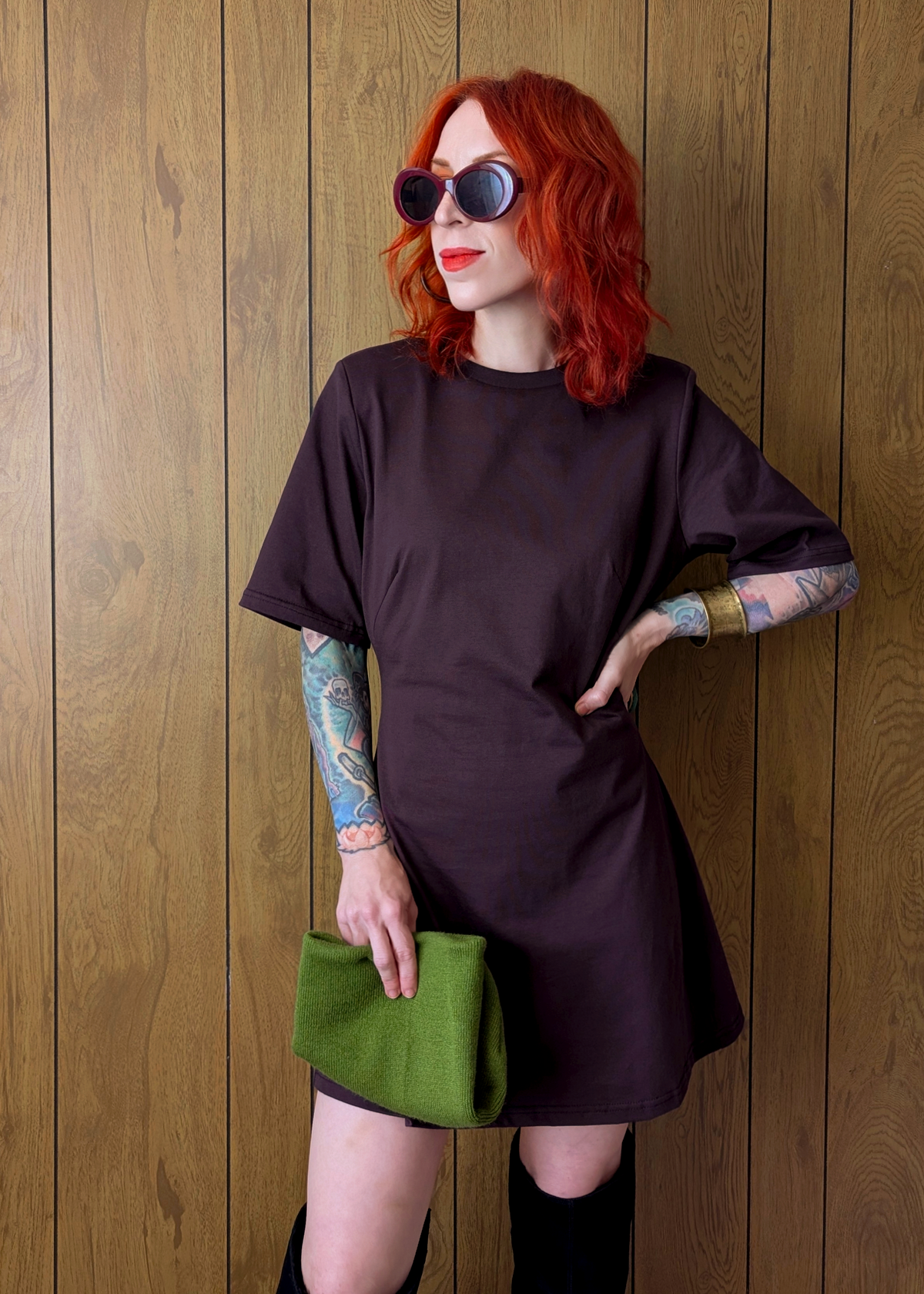 Person wearing a dark plum eggplant dress with a green clutch against a wooden wall