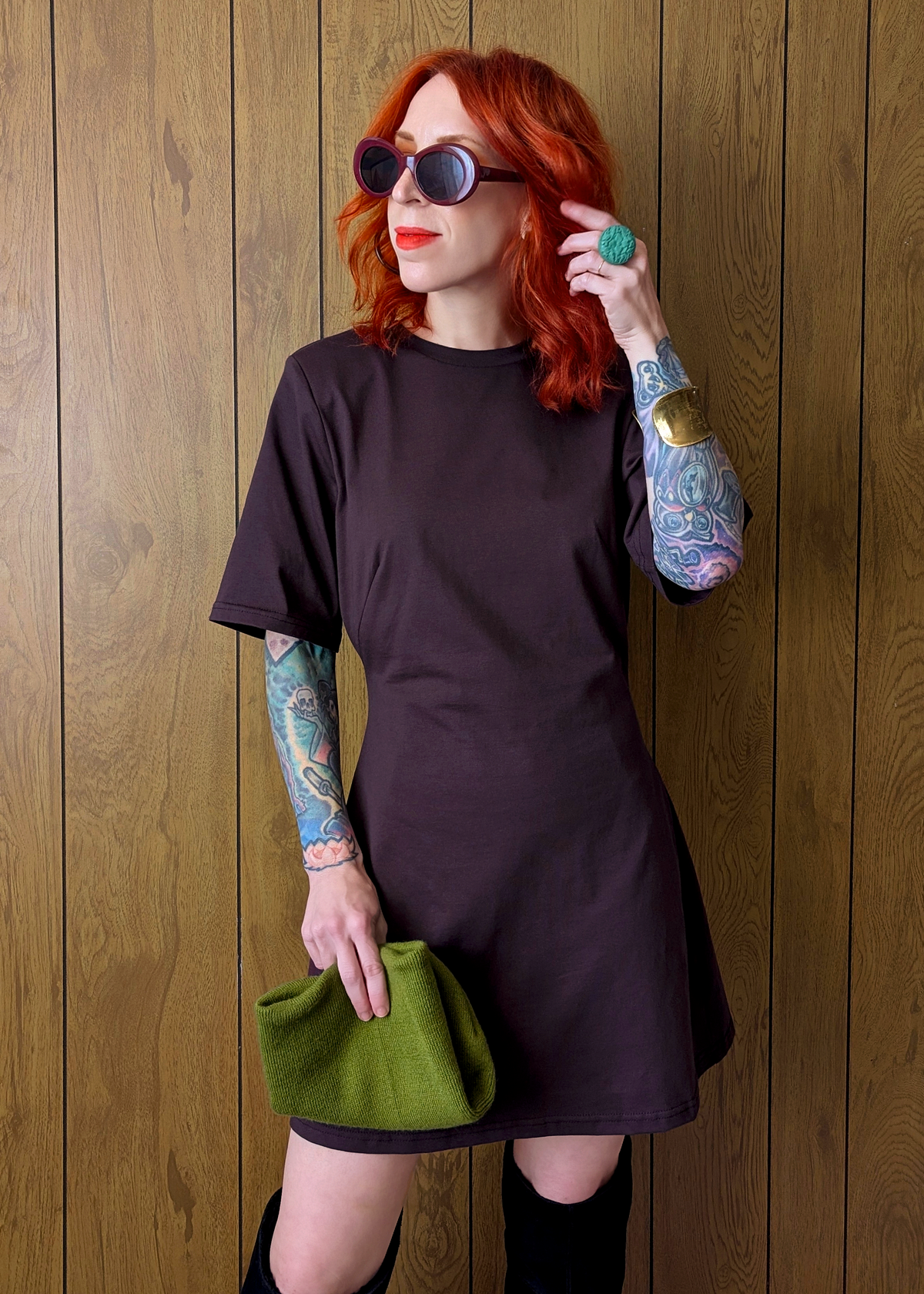 Woman with red hair wearing a plum / eggplant colored t-shirt dress and sunglasses, standing against a wooden wall.