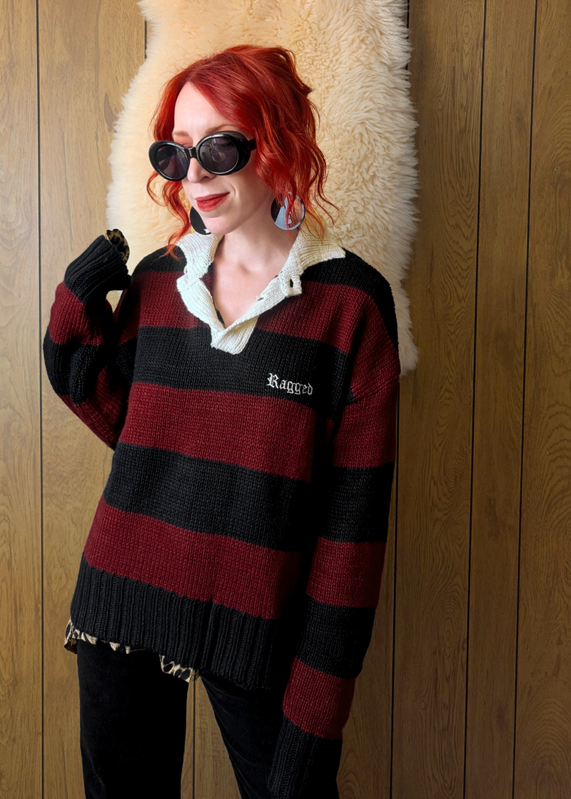 Person wearing a black and burgundy striped polo  sweater with 'Ragged' embroidery at chest, standing against a wooden wall.