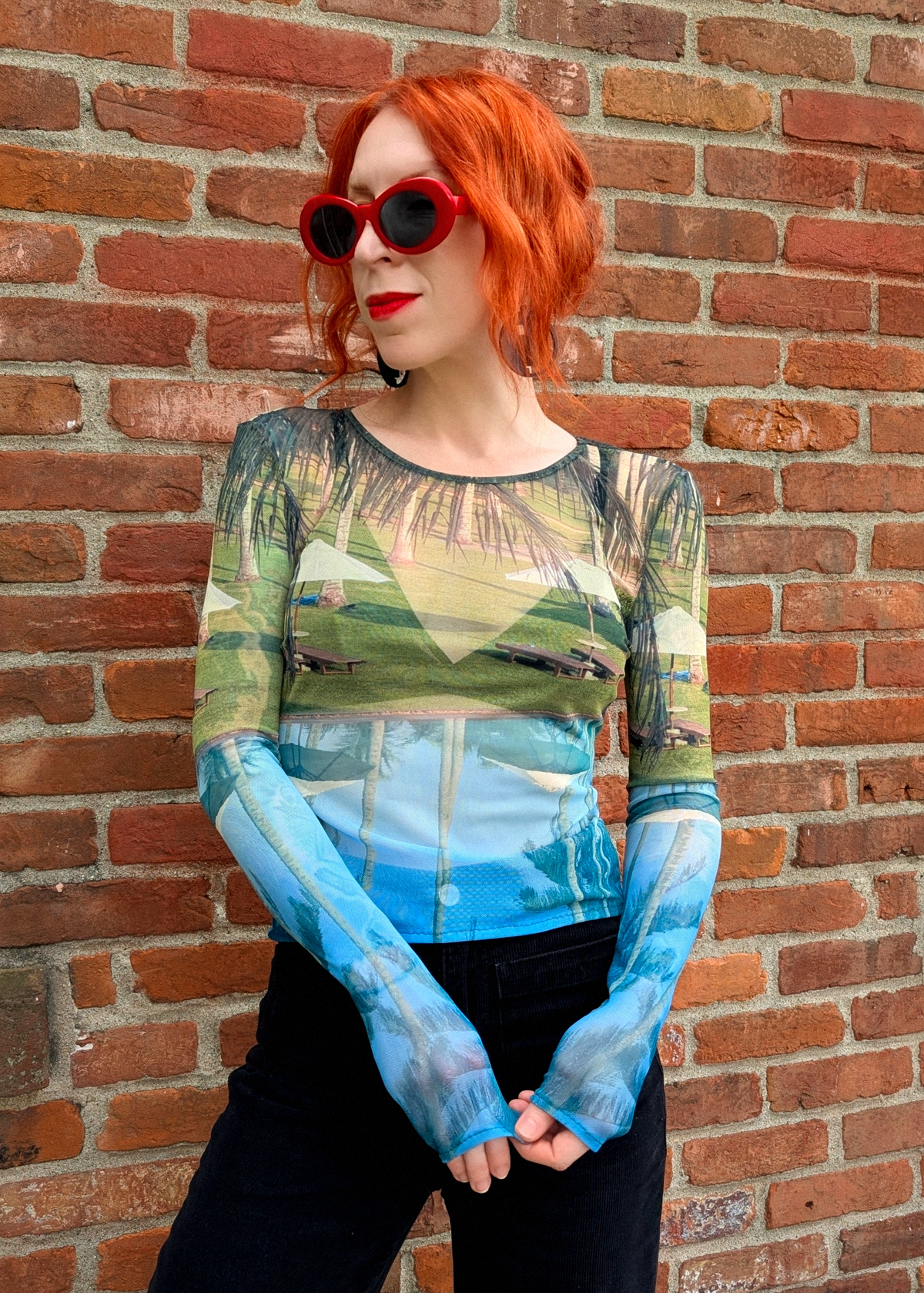 Person with red hair wearing a patterned top and sunglasses against a brick wall.