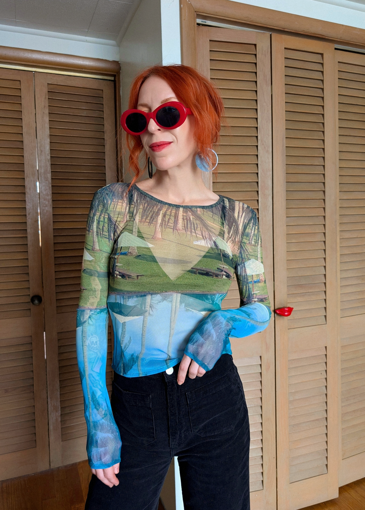 Person wearing a tropical patterned long sleeve mesh top with a scenic design, standing in front of wooden shutters.