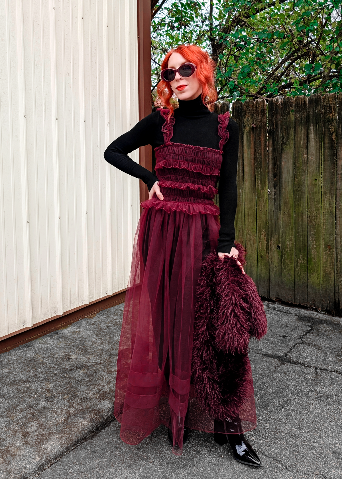 Woman in a sheer burgundy tulle maxi dress with ruffles and a black top standing outdoors.