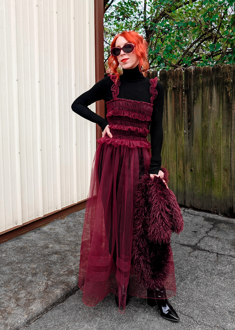 Woman in a sheer burgundy tulle maxi dress with ruffles and a black top standing outdoors.