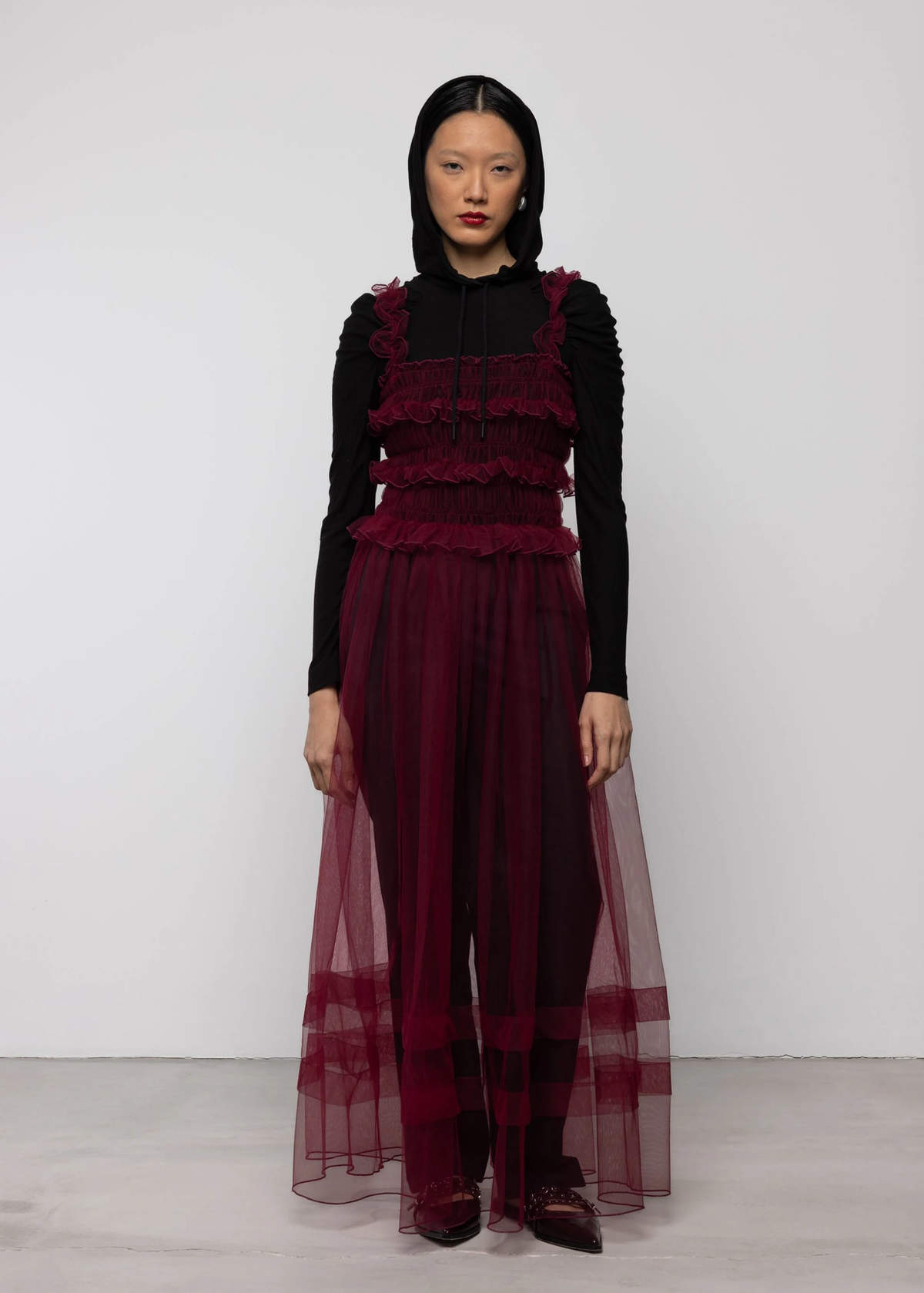 Woman wearing a sheer burgundy tulle dress over a black top and black trousers against a white background