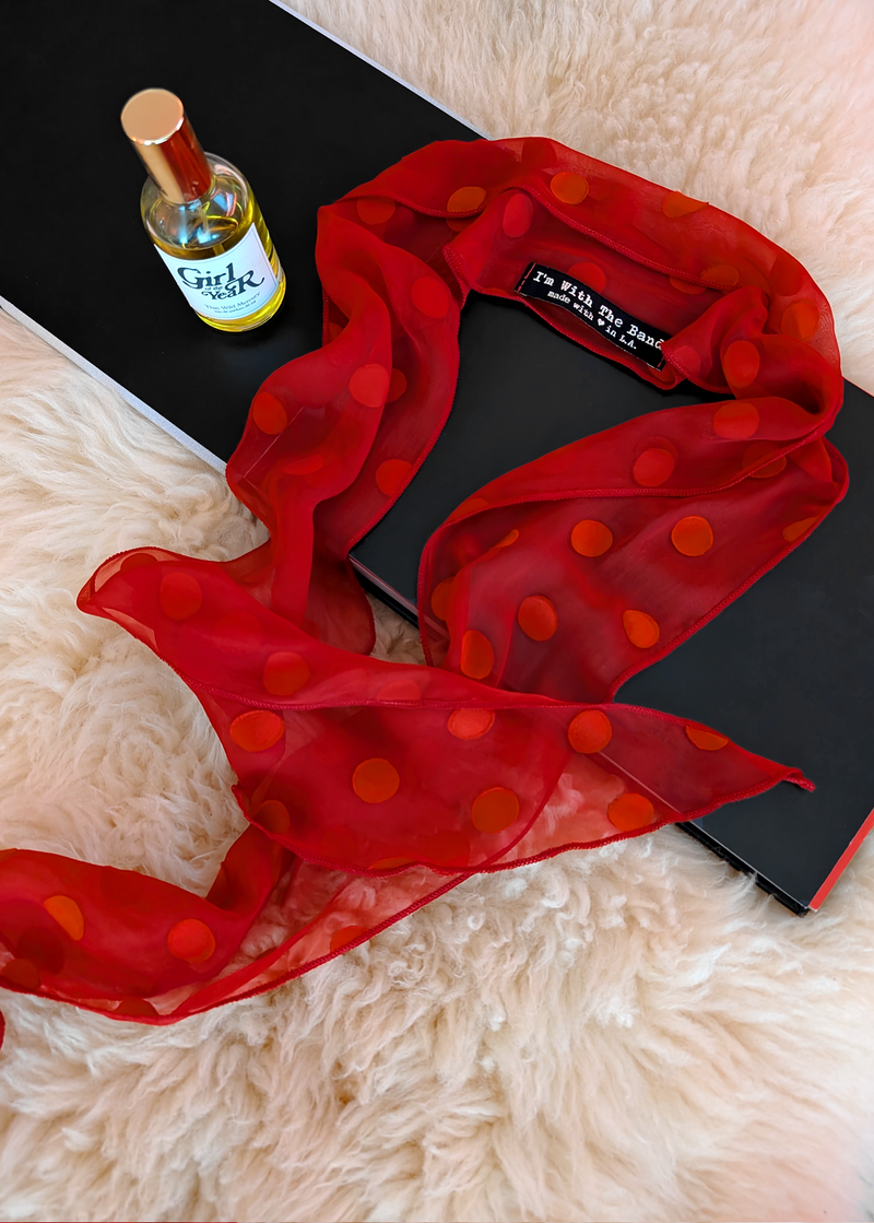 Lifestyle photo of a red polka dot scarf laid flat on a cream faux fur rug with a black book and bottle of perfume