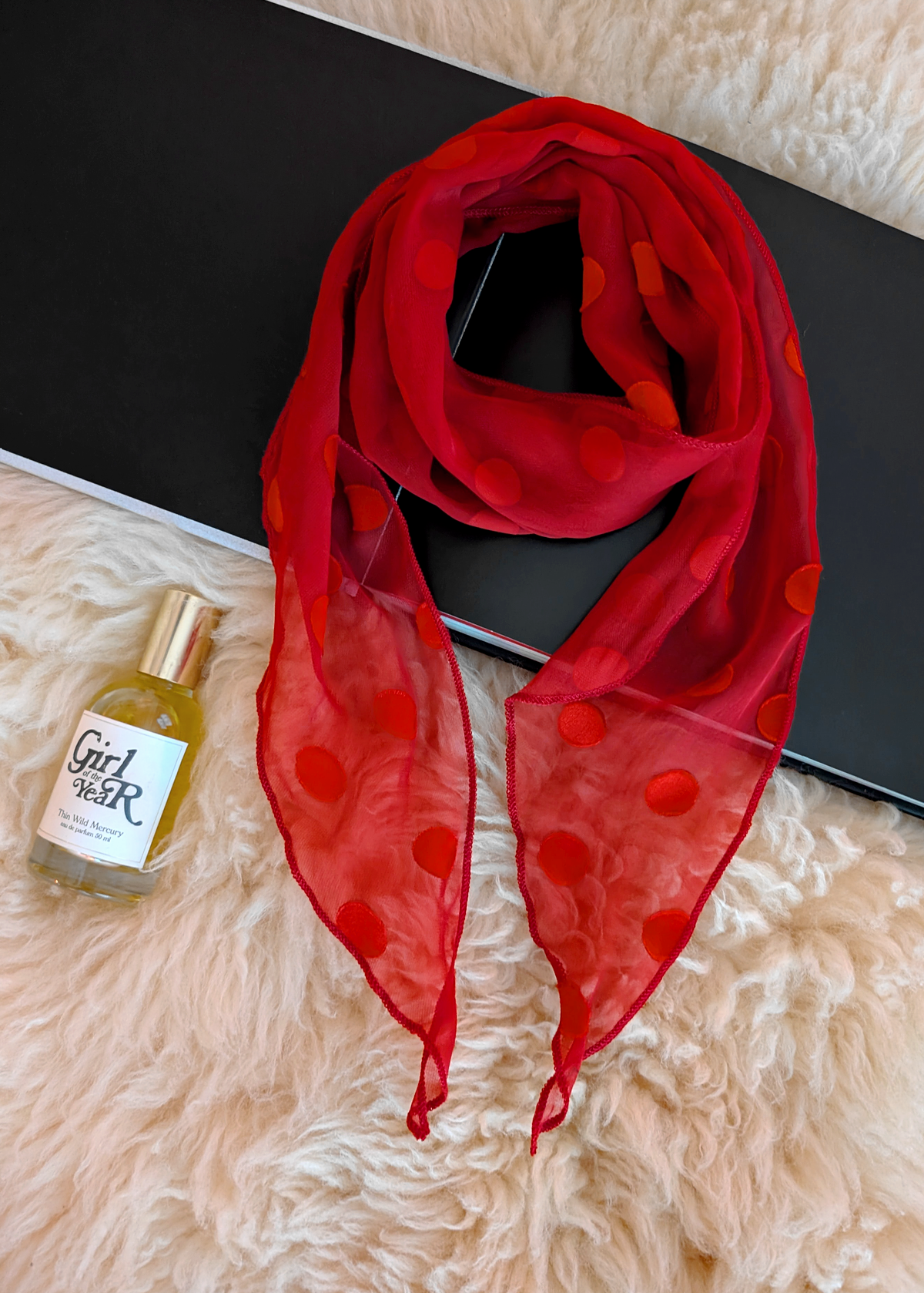 Lifestyle photo of a red polka dot scarf laid flat on a cream faux fur rug with a black book and bottle of perfume