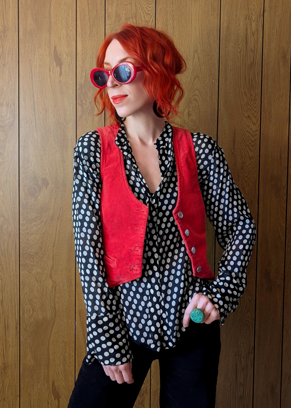 Woman with red hair wearing a polka dot blouse and red vest against a wooden wall.