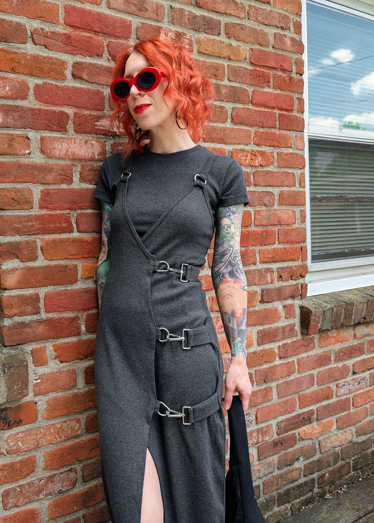 Person wearing a charcoal grey rib tank dress with silver buckles, layered over a charcoal grey rib tee, posed against a brick wall background