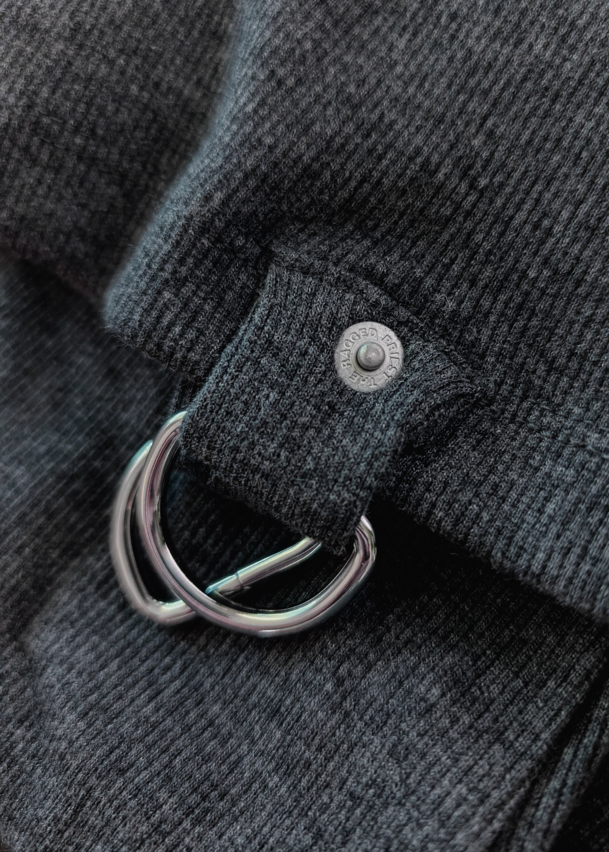 Close-up of a dark gray fabric with a metal D-ring and rivet with "ragged priest" engraving