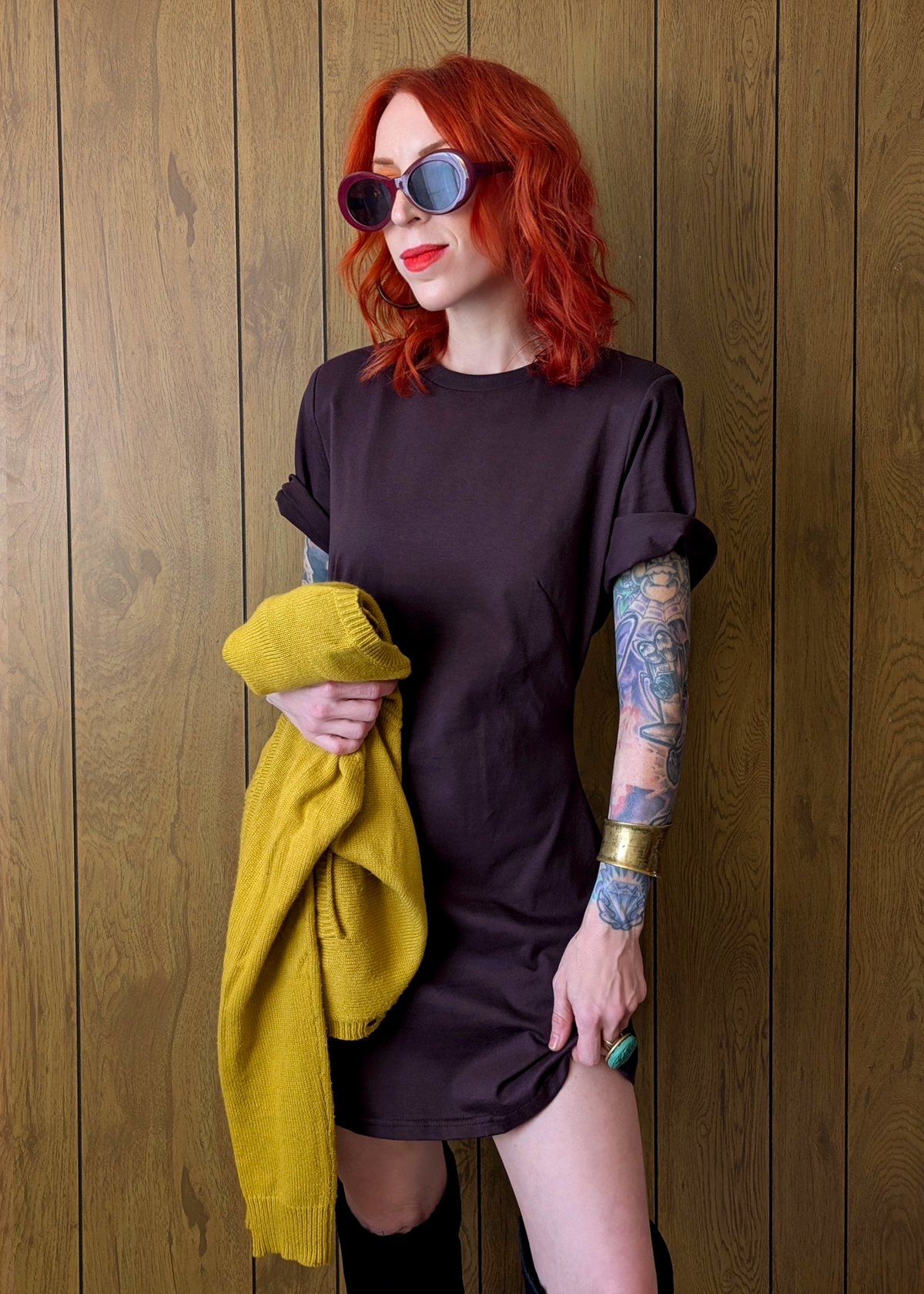 Woman with red hair and tattoos wearing a dark plum t-shirt dress against a wooden wall.