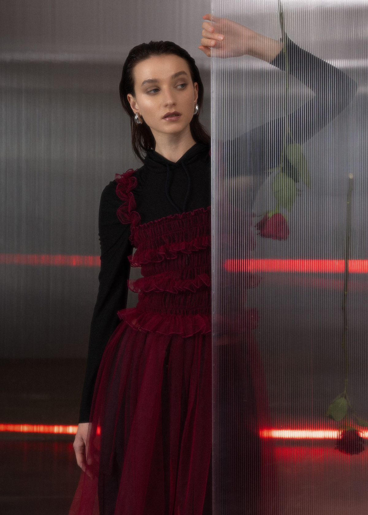 Woman wearing a black top under a sheer ruffled tulle burgundy dress holding a rose in a dark, moody setting with red lighting.
