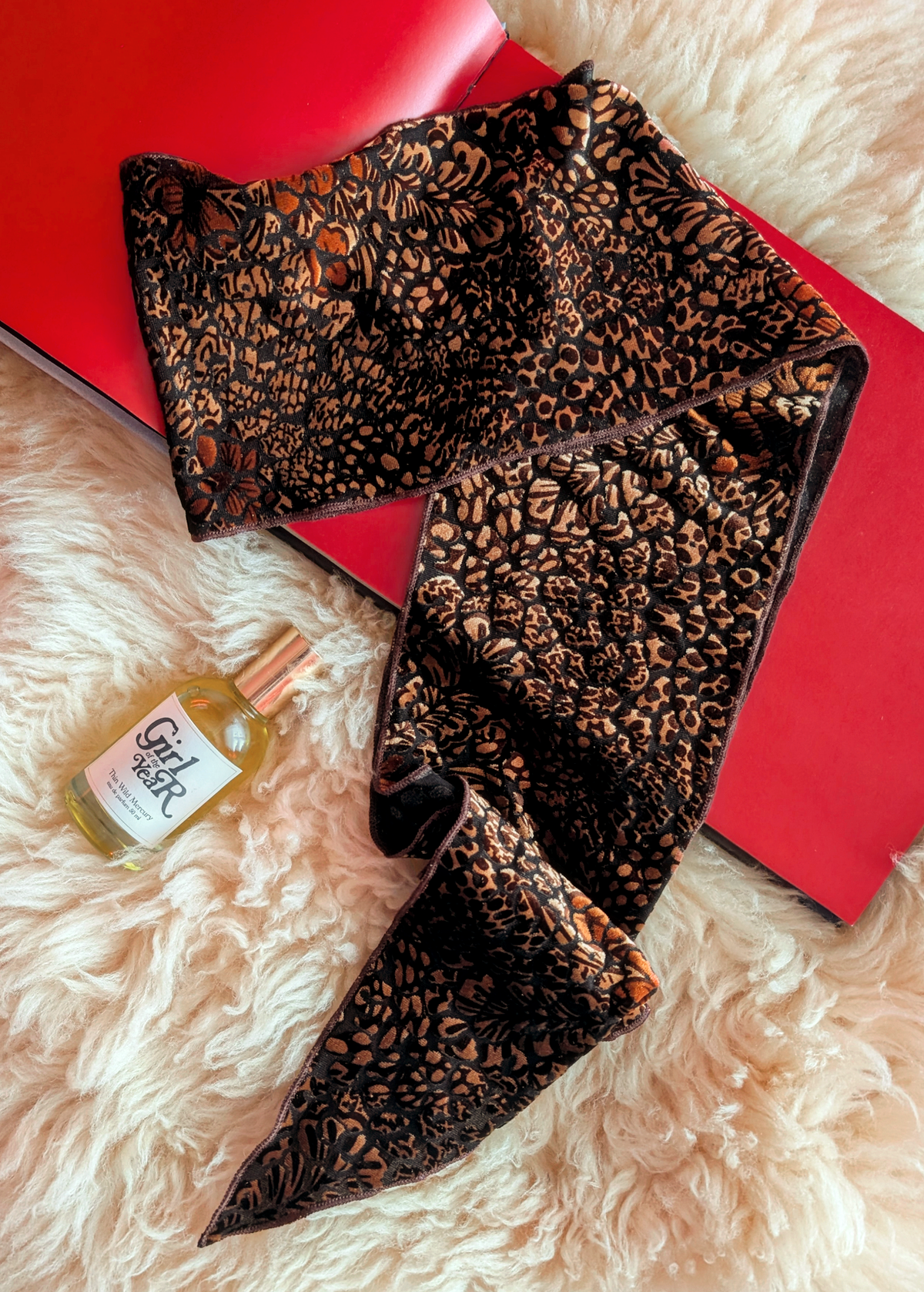 Lifestyle photo of a animal print burnout velvet scarf laid flat on a cream faux fur rug with a red book and bottle of perfume