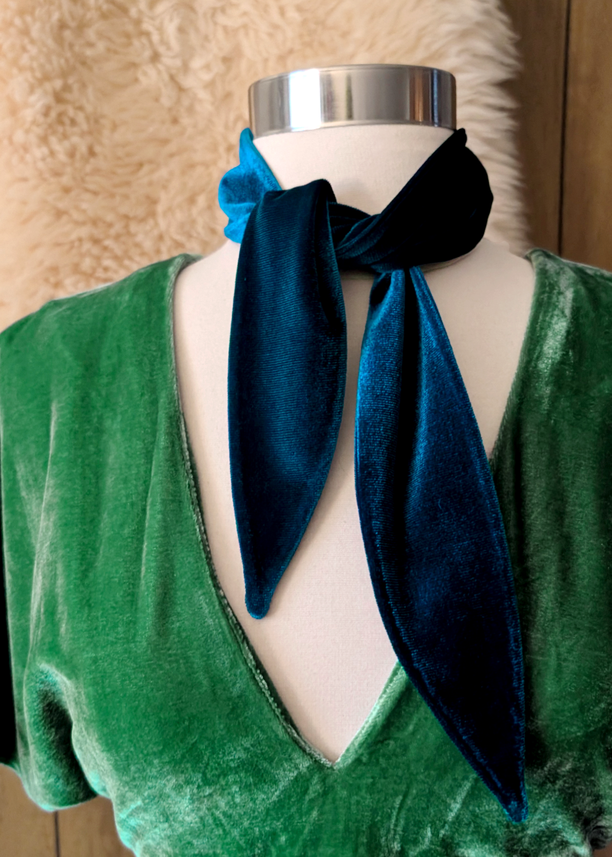 Mannequin wearing a green velvet dress with a blue scarf around its neck against a textured beige wall.