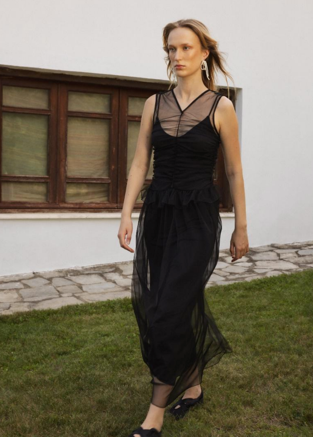 Woman wearing a black tulle dress standing outdoors on grass with a white wall and window in the background.