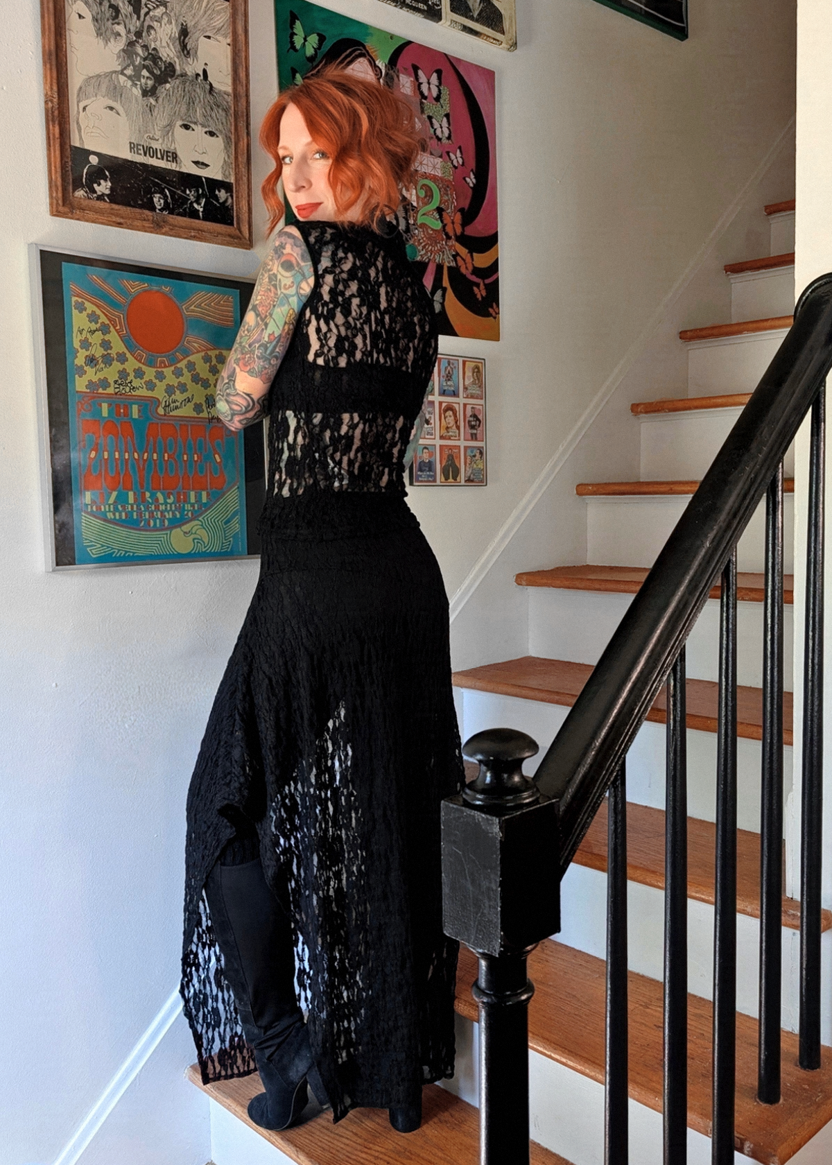 A woman with red hair and tattoos stands on wooden stairs wearing a black lace top and skirt, over black leggings and boots, with framed posters on the wall behind her.