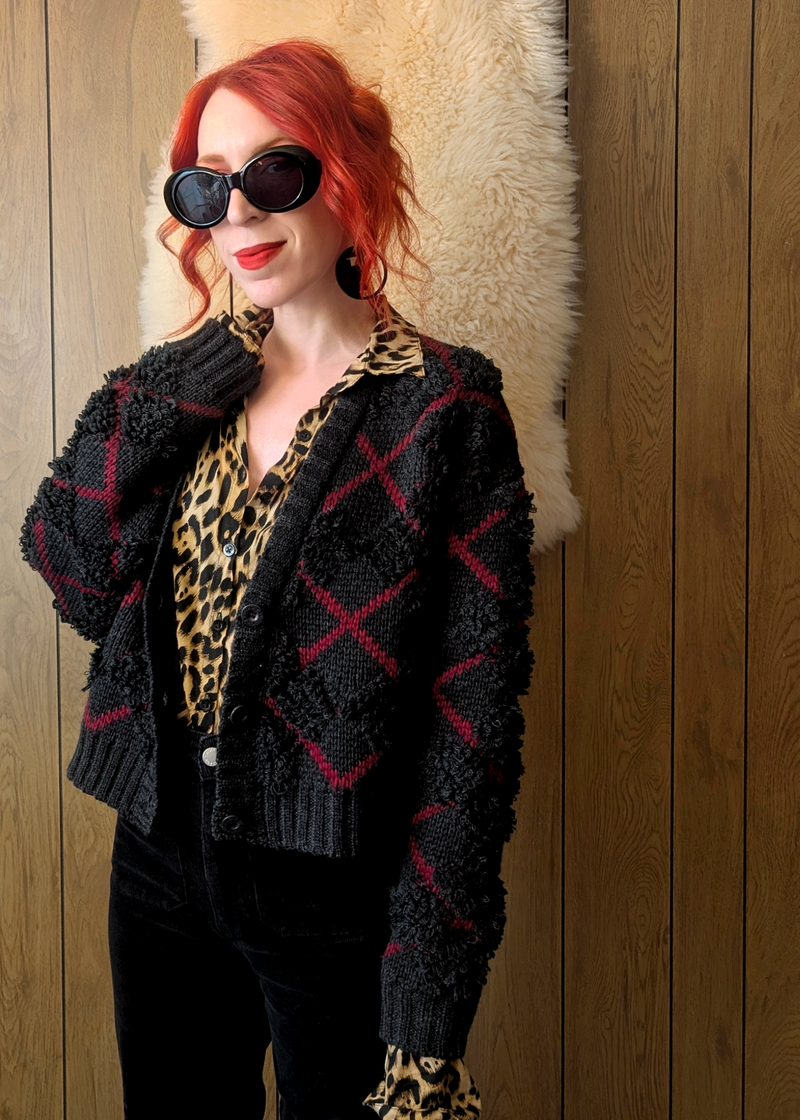 Person wearing a charcoal and red patterned cardigan over a leopard print shirt against a wooden wall.