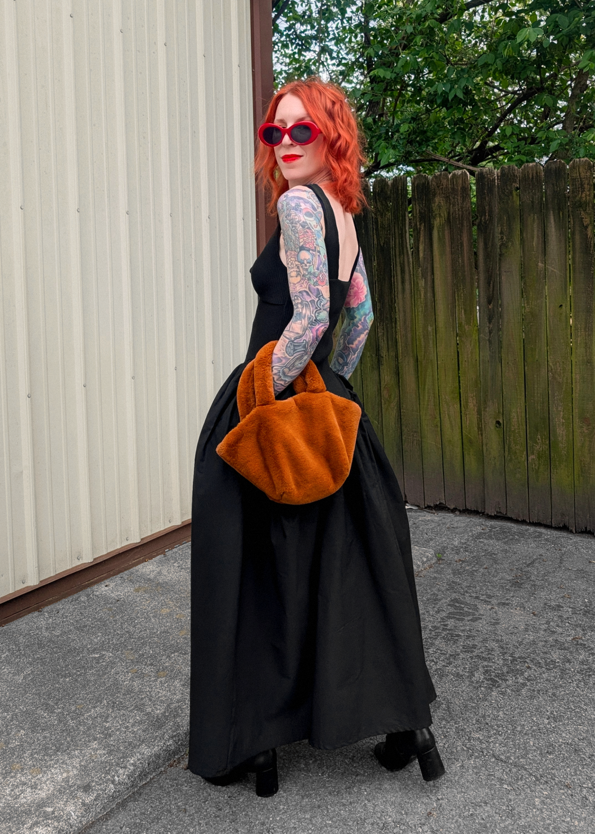 Person with red hair and tattoos wearing a black dress and holding an orange bag outdoors.