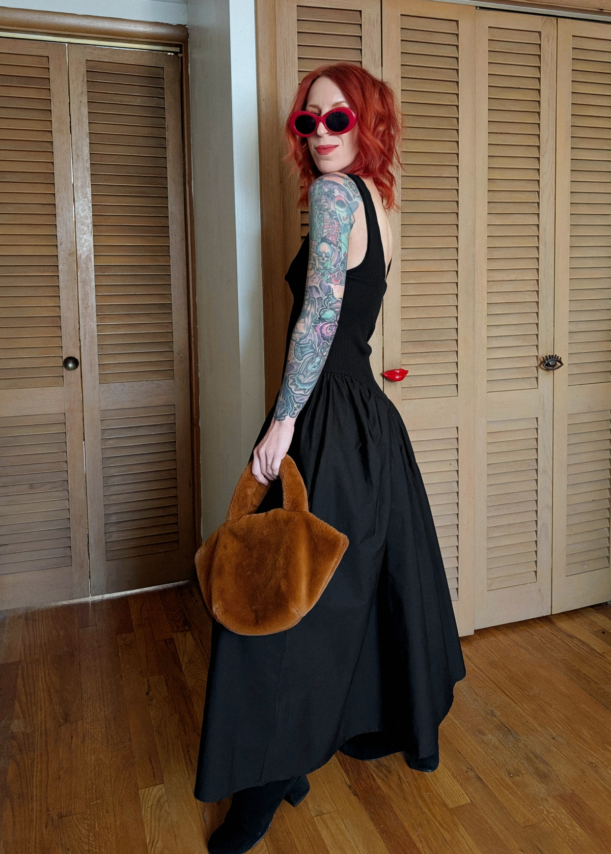 Woman in a black dress holding a n rust faux fur bag in a room with wooden floors and shutters.