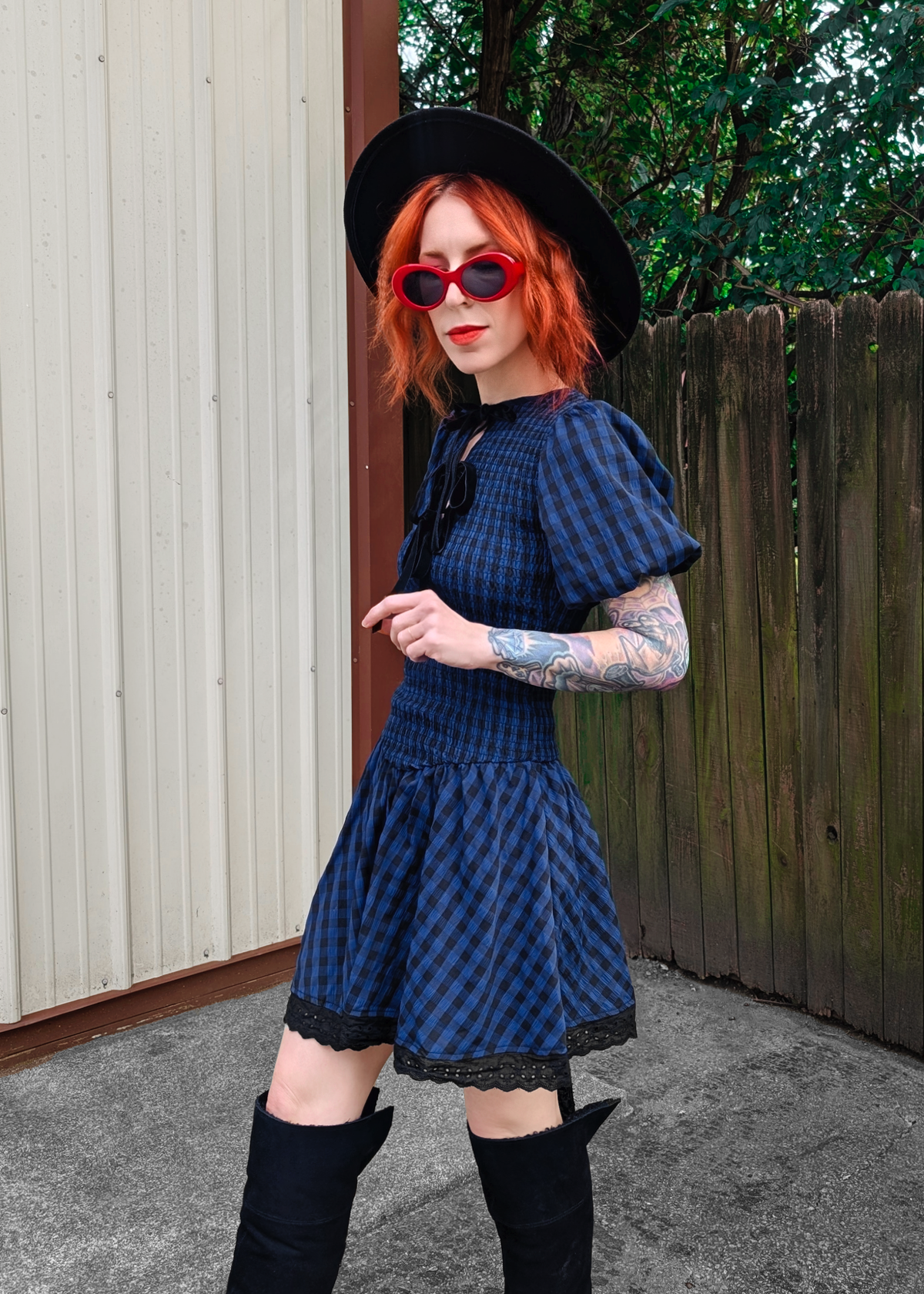 Person wearing a blue and black checkered dress, black boots, and a black hat outdoors.