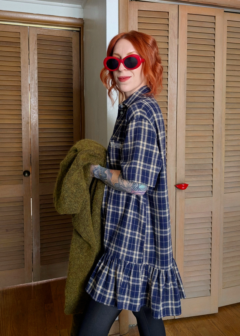 Woman in a blue plaid dress and red sunglasses standing in a room with wooden shutters.