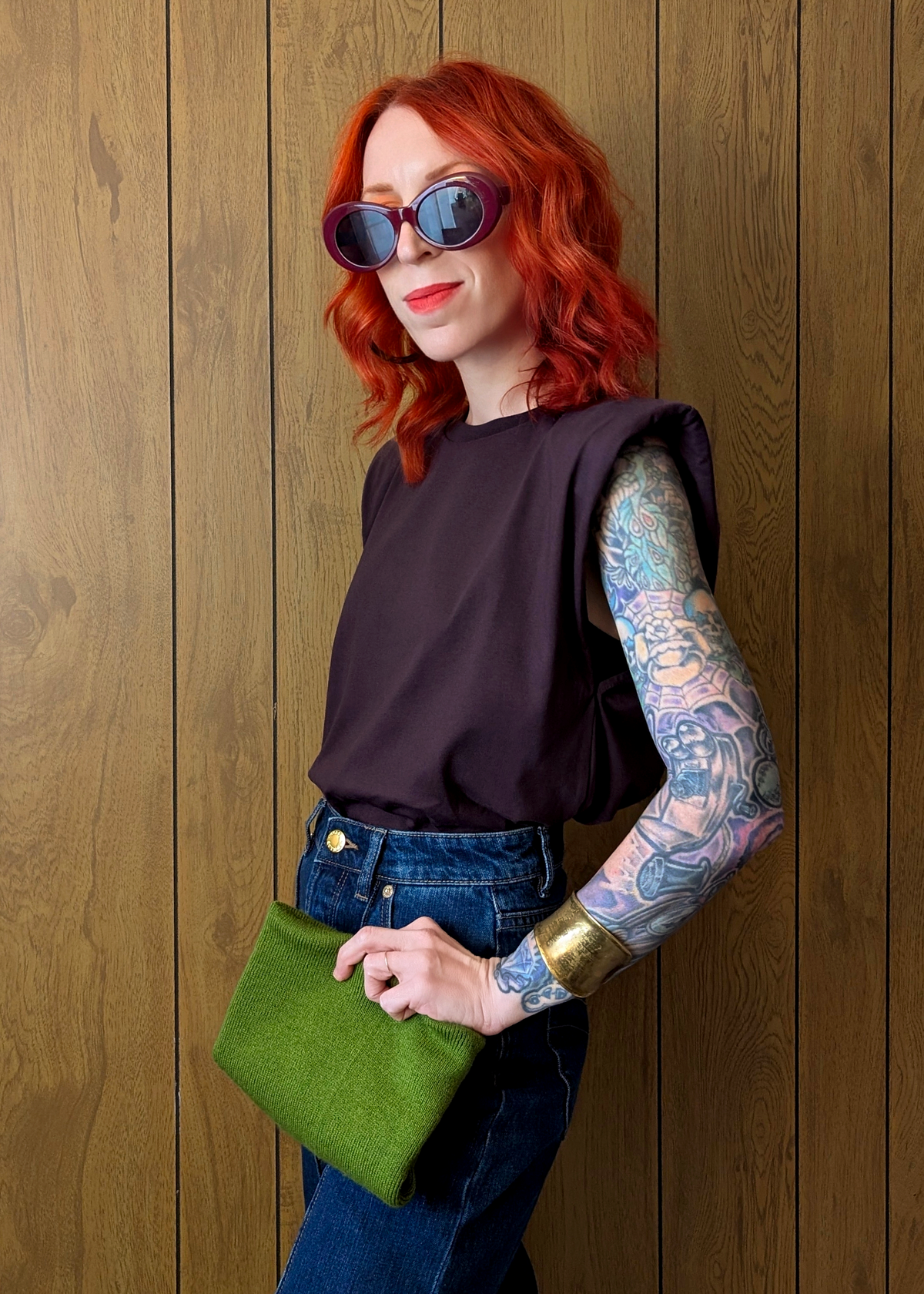Woman with red hair and tattoos waring a sleeveless plum t-shirt holding a green clutch against a wooden wall