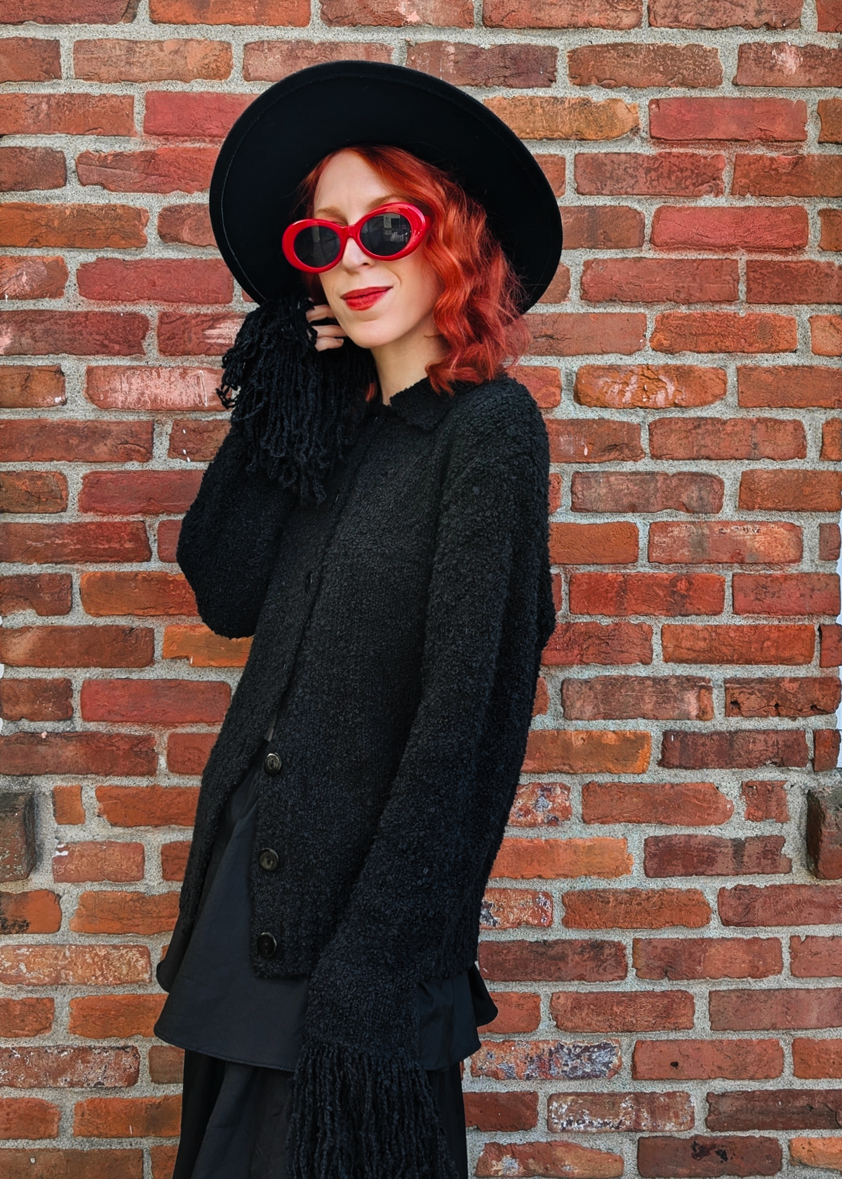 Person wearing a black textured cardigan, black hat, and red sunglasses against a brick wall.
