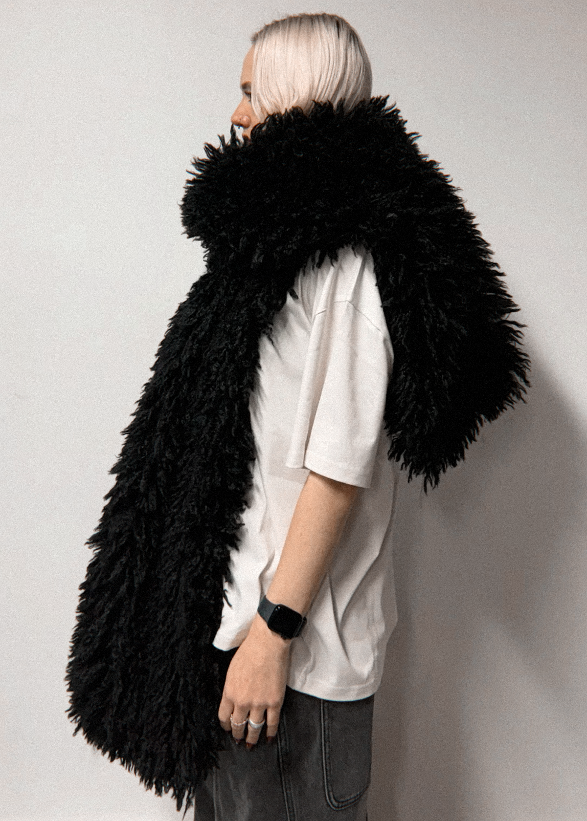 A person with platinum blonde hair is shown from the side, wearing a voluminous, long-pile black faux fur scarf over a white t-shirt and dark gray jeans, standing against a plain light gray wall.