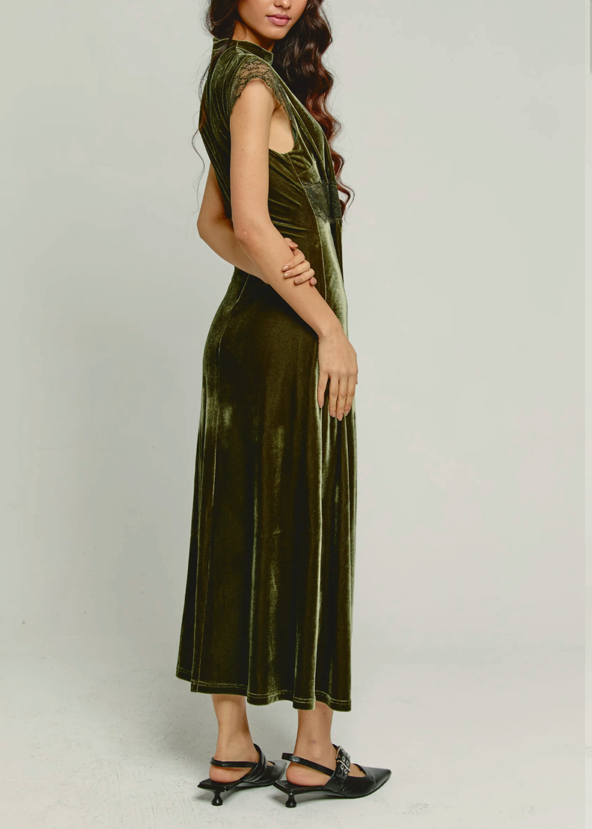 A woman modeling a sleeveless olive green velvet midi dress with lace capped sleeves, paired with black pointed-toe flats, against a plain light gray studio background.