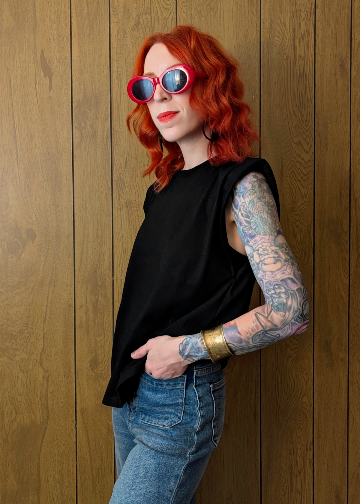 Person with red hair and tattoos wearing sunglasses and a black top against a wooden wall.