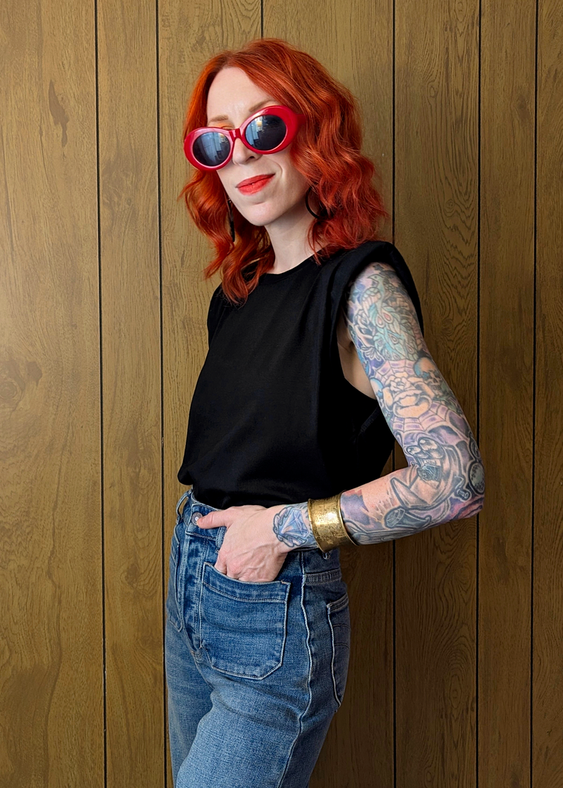 Person with red hair and tattoos wearing sunglasses and a black sleeveless top against a wooden wall.