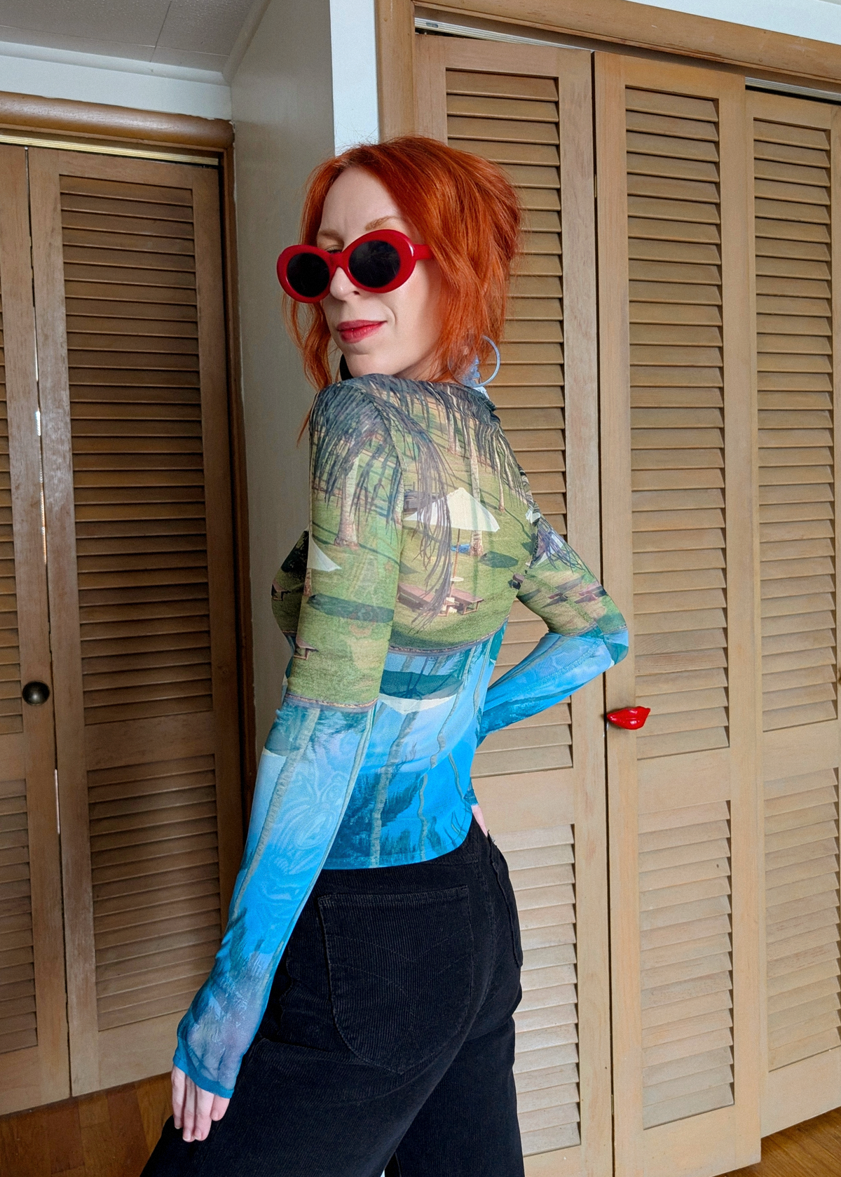 Person with red hair wearing sunglasses and a patterned mesh top in front of wooden shutters.