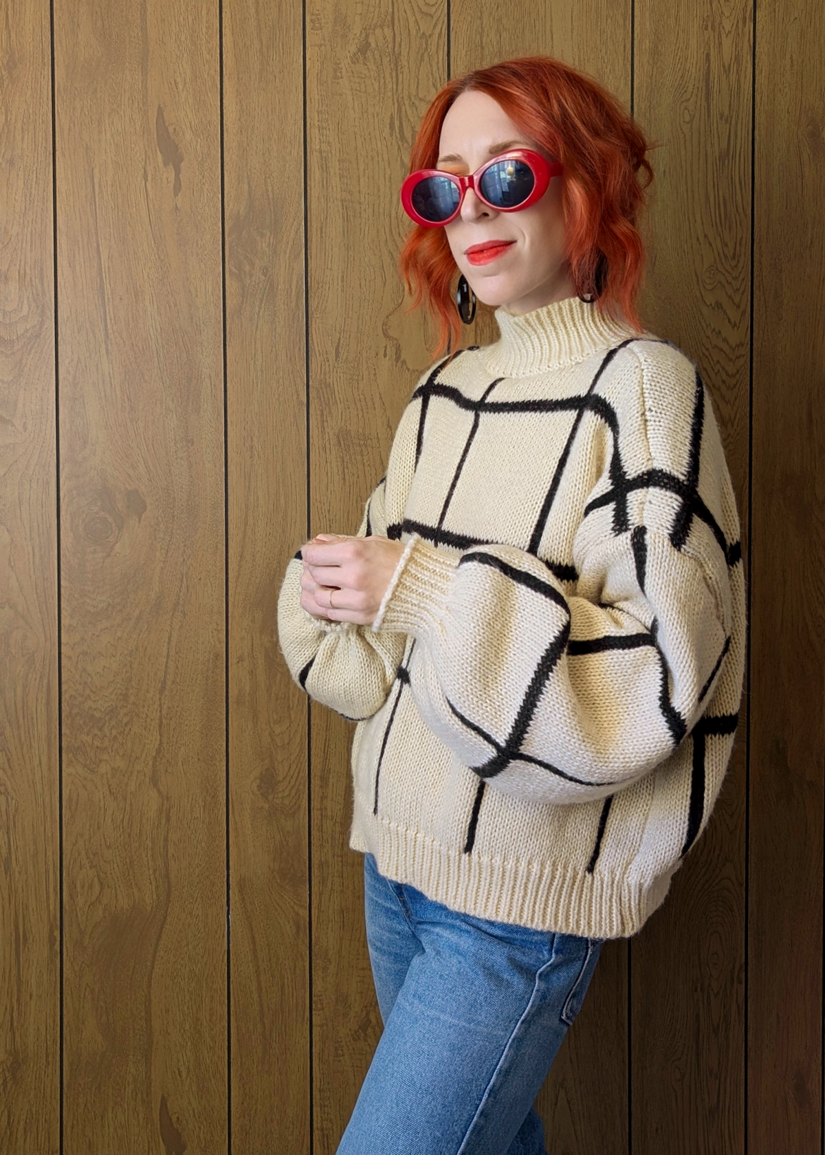 Person wearing a beige sweater with black abstract patterns, blue jeans, and red sunglasses against a wooden panel background.