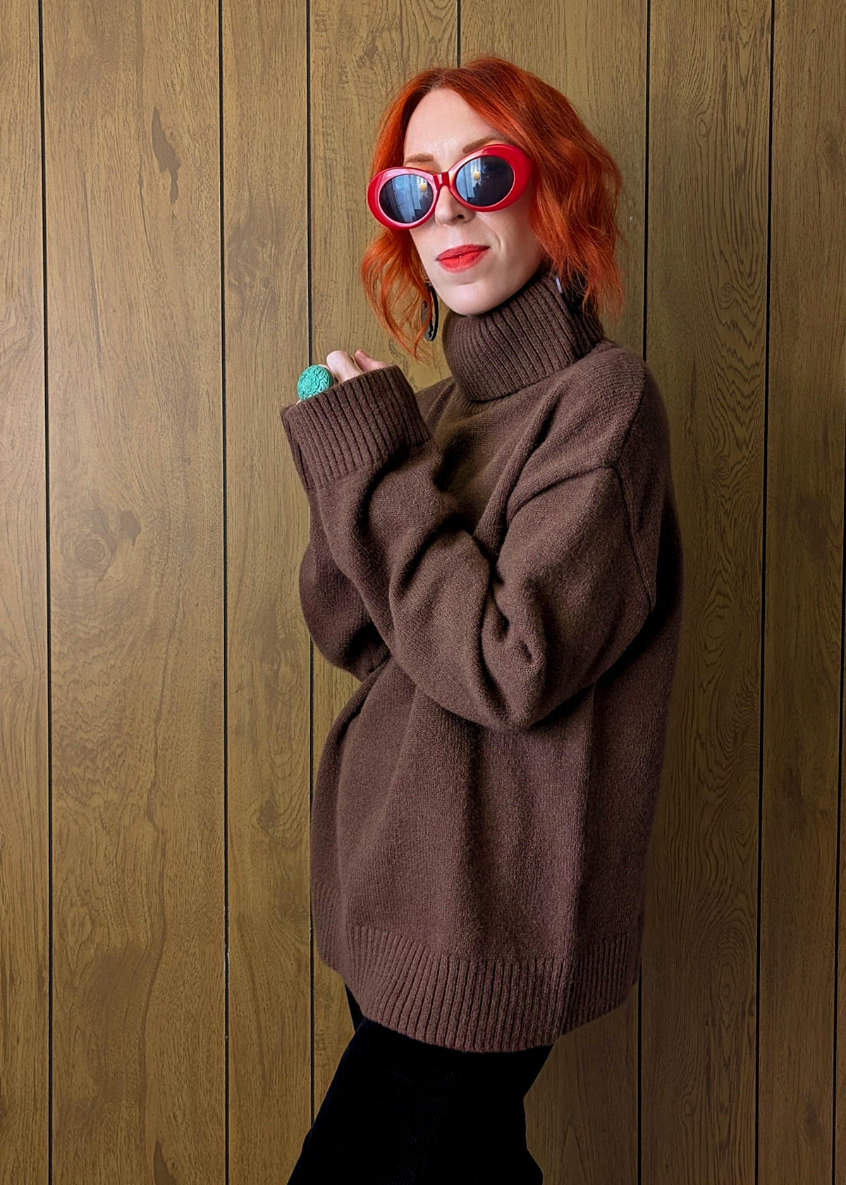 Person wearing a brown sweater and red sunglasses against a wooden wall