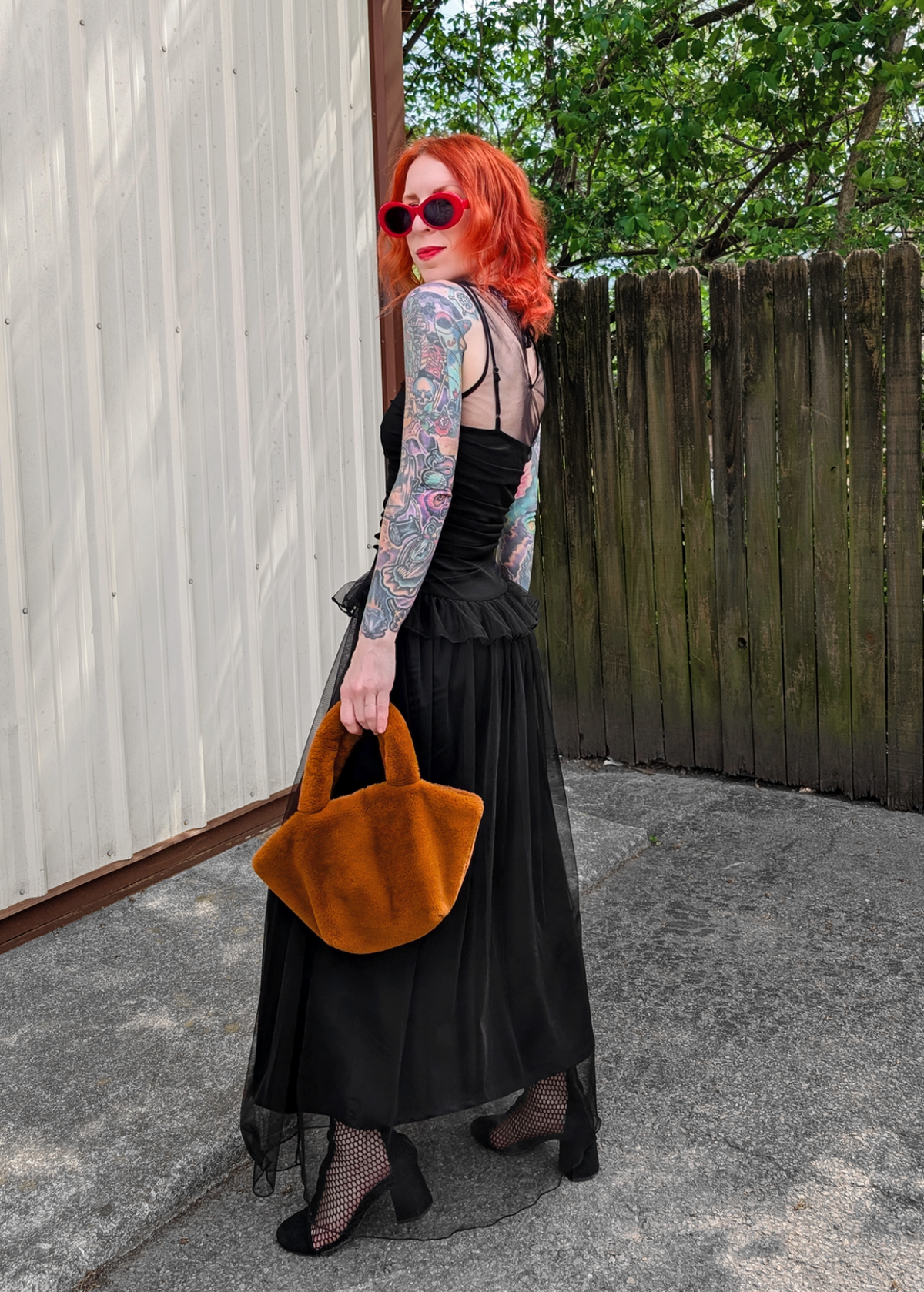 Person with red hair and tattoos wearing a black tulle dress and holding an faux  fur handbag against a wooden fence outside. 