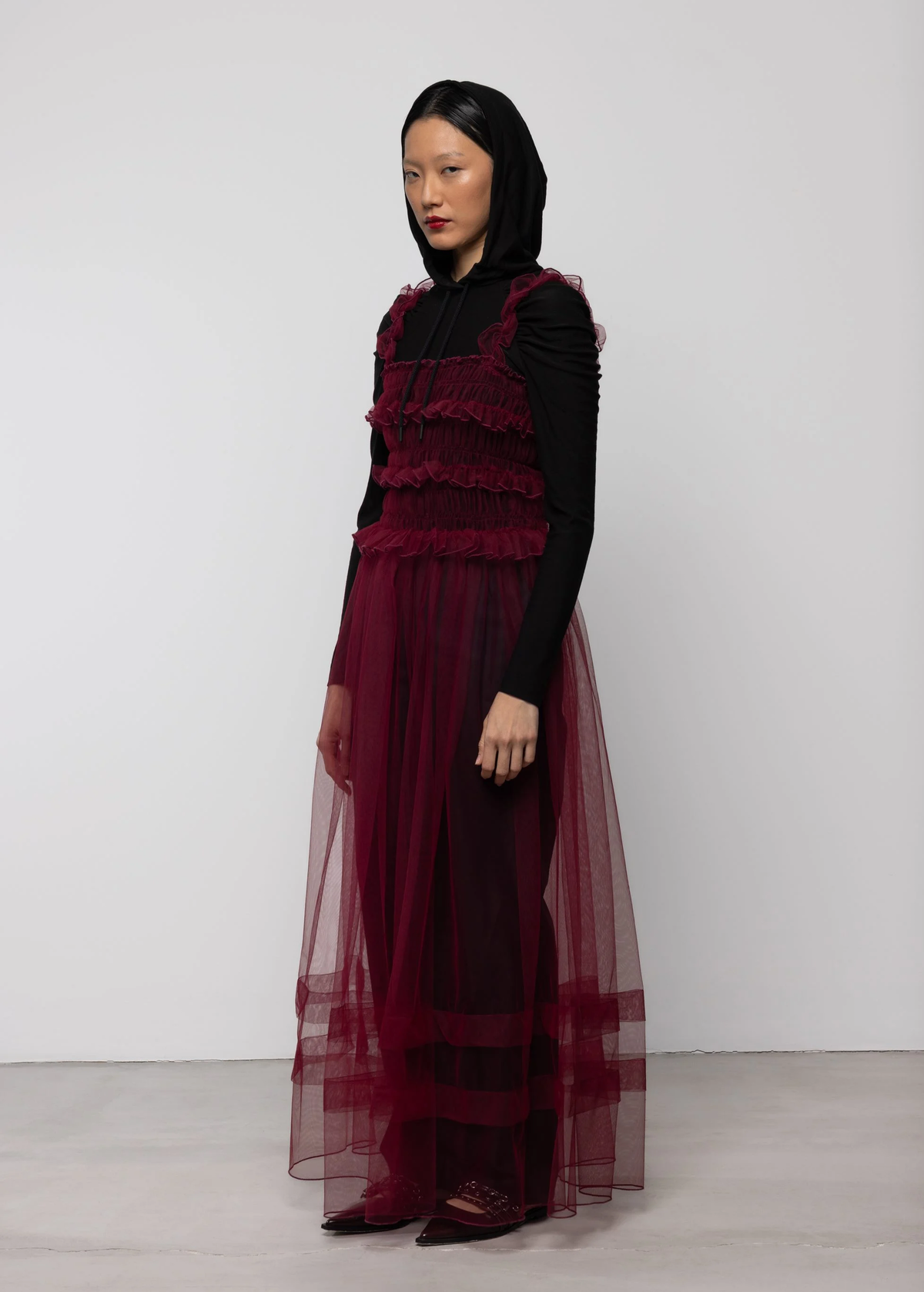 Woman wearing a sheer burgundy tulle dress over a black top and black trousers against a white background