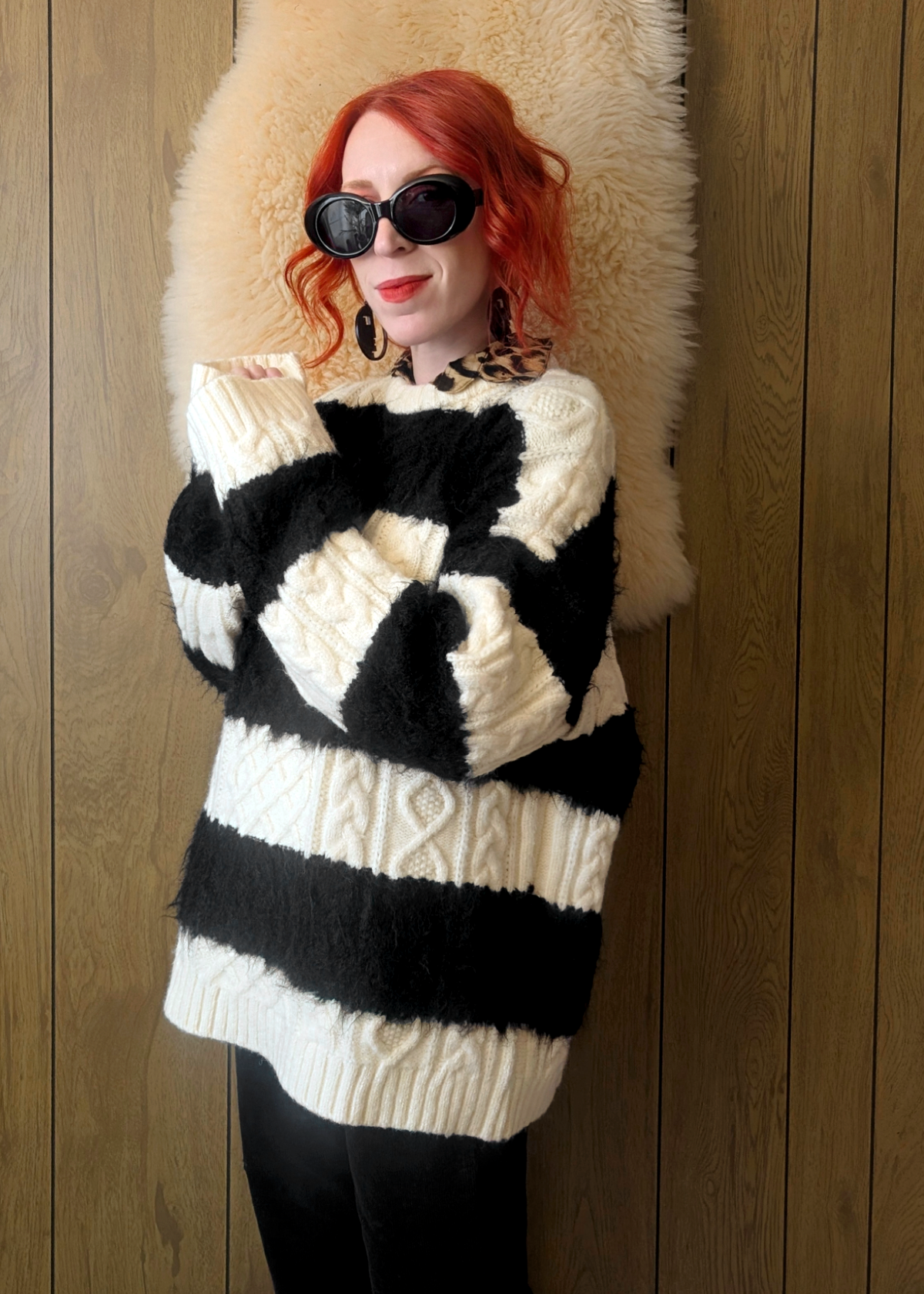 Person wearing a black and cream striped cable knit sweater layered over a leopard print collared shirt, posed against a wooden wall.