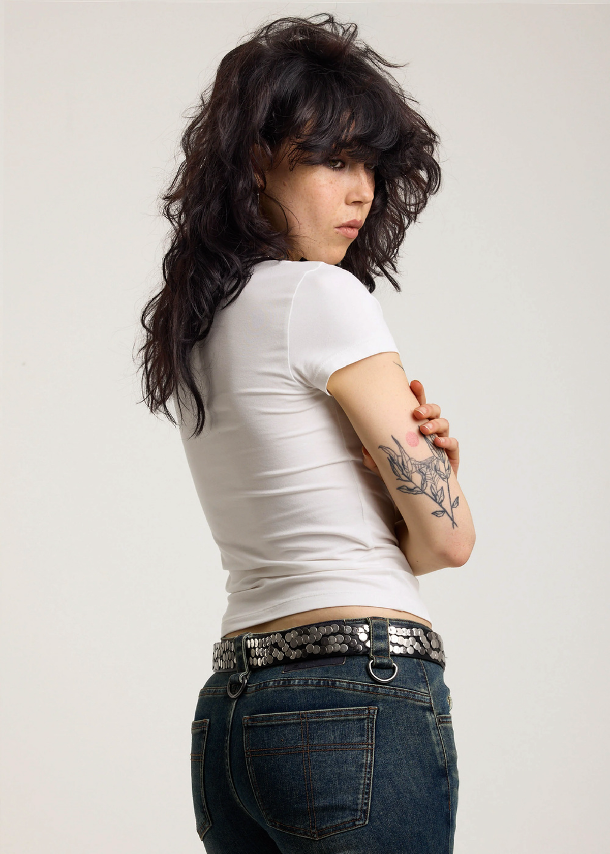 Side view of a woman wearing an ivory baby tee and blue jeans with a plain background