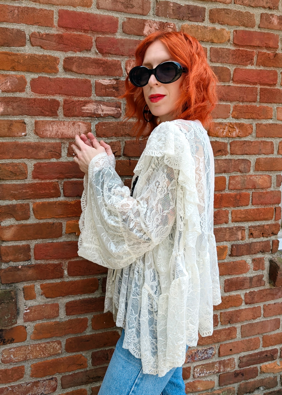 A woman with vibrant red hair and black sunglasses leans against a red brick wall, wearing an ivory lace long-sleeve blouse and blue jeans.